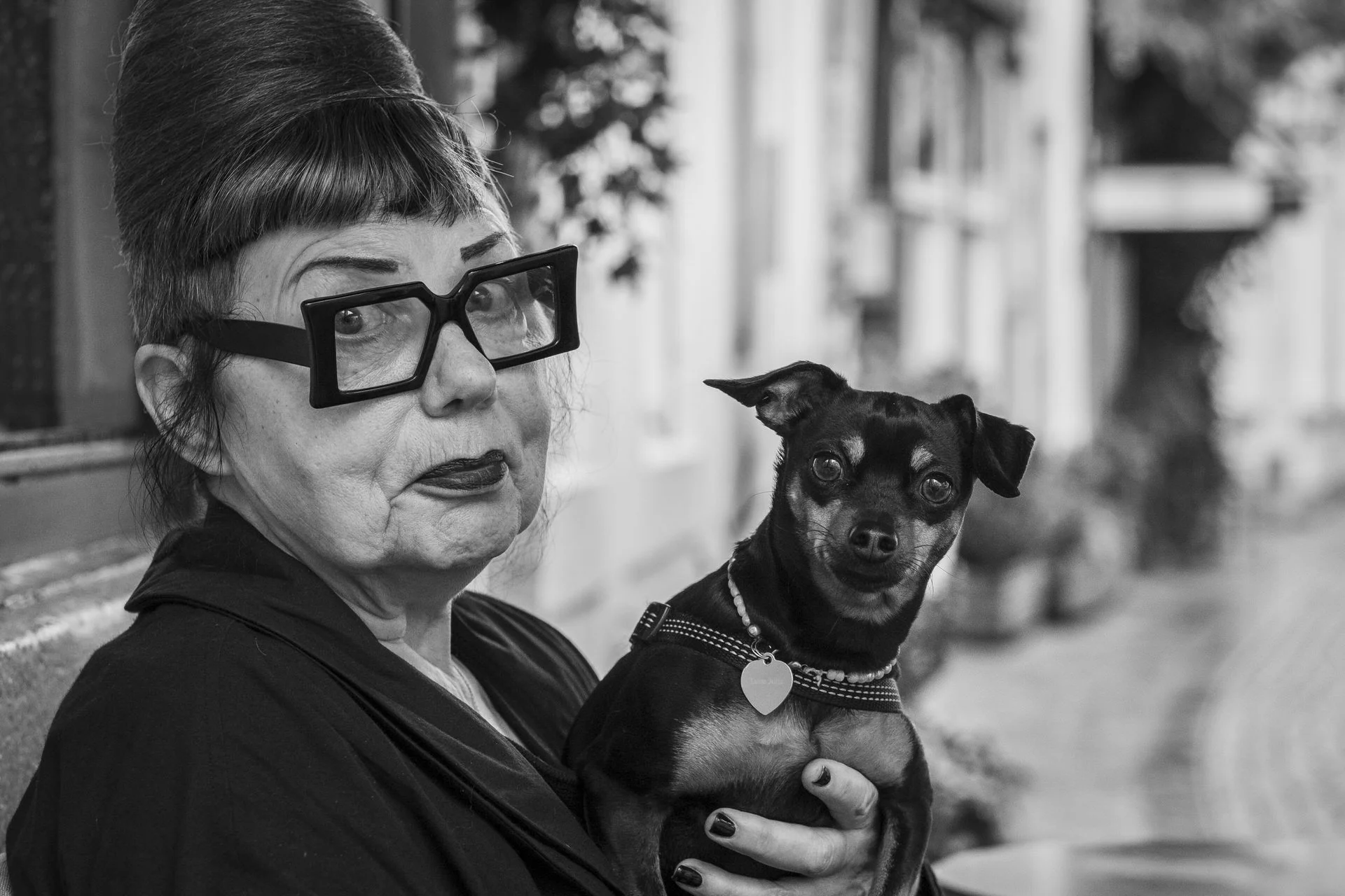A woman with short, styled hair and large glasses holds a small, dark-colored dog with a striped collar and a heart-shaped tag, both seated outdoors.