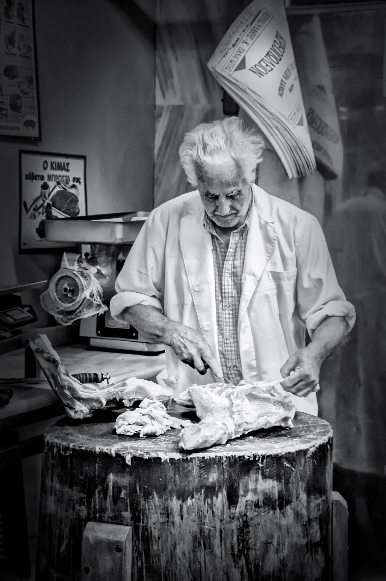 An elderly man with white hair is cutting meat on a wooden table in a butcher shop, with various tools and posters visible in the background.