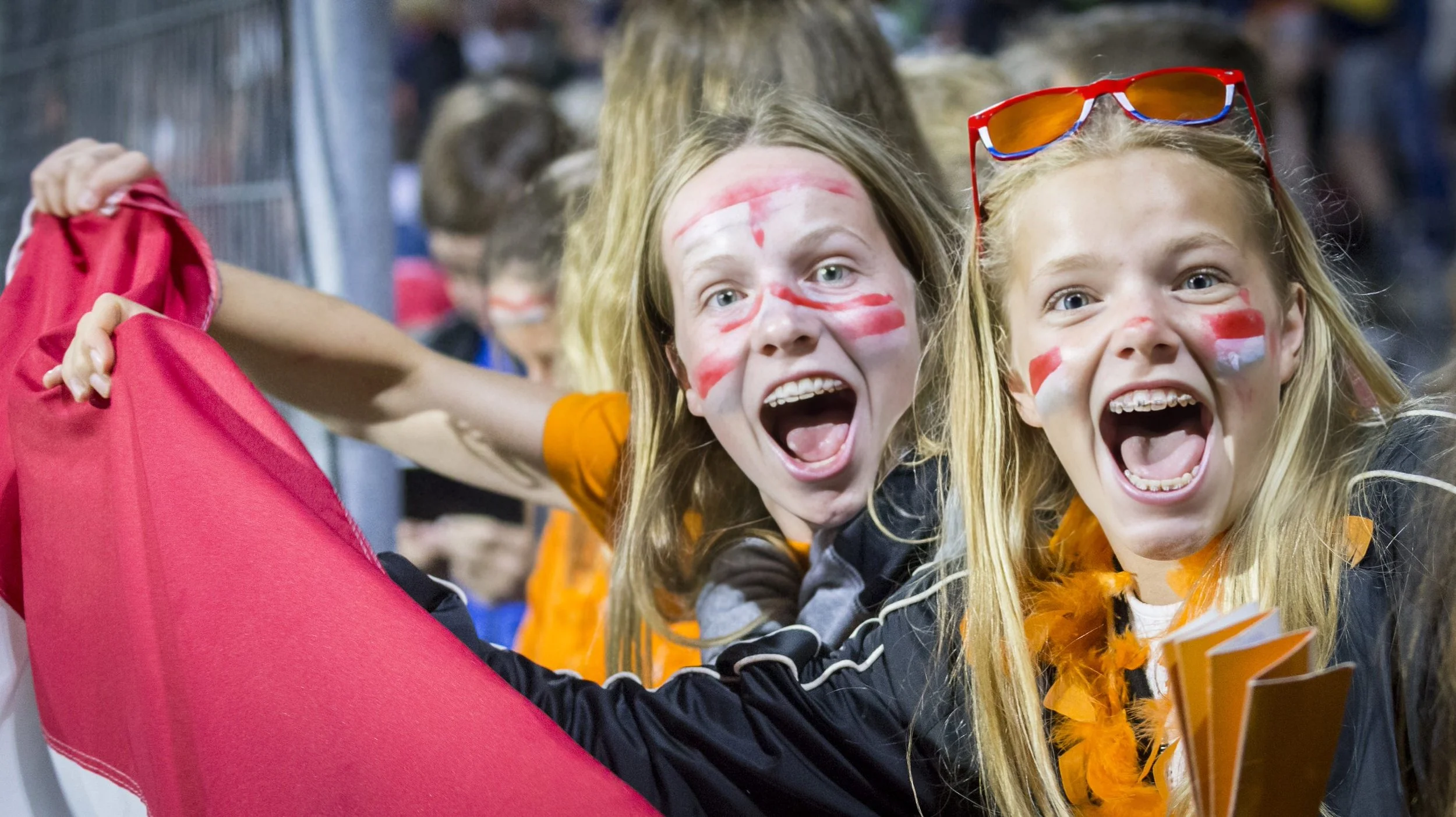 Two excited young girls with face paint and sunglasses celebrating at a crowded event.