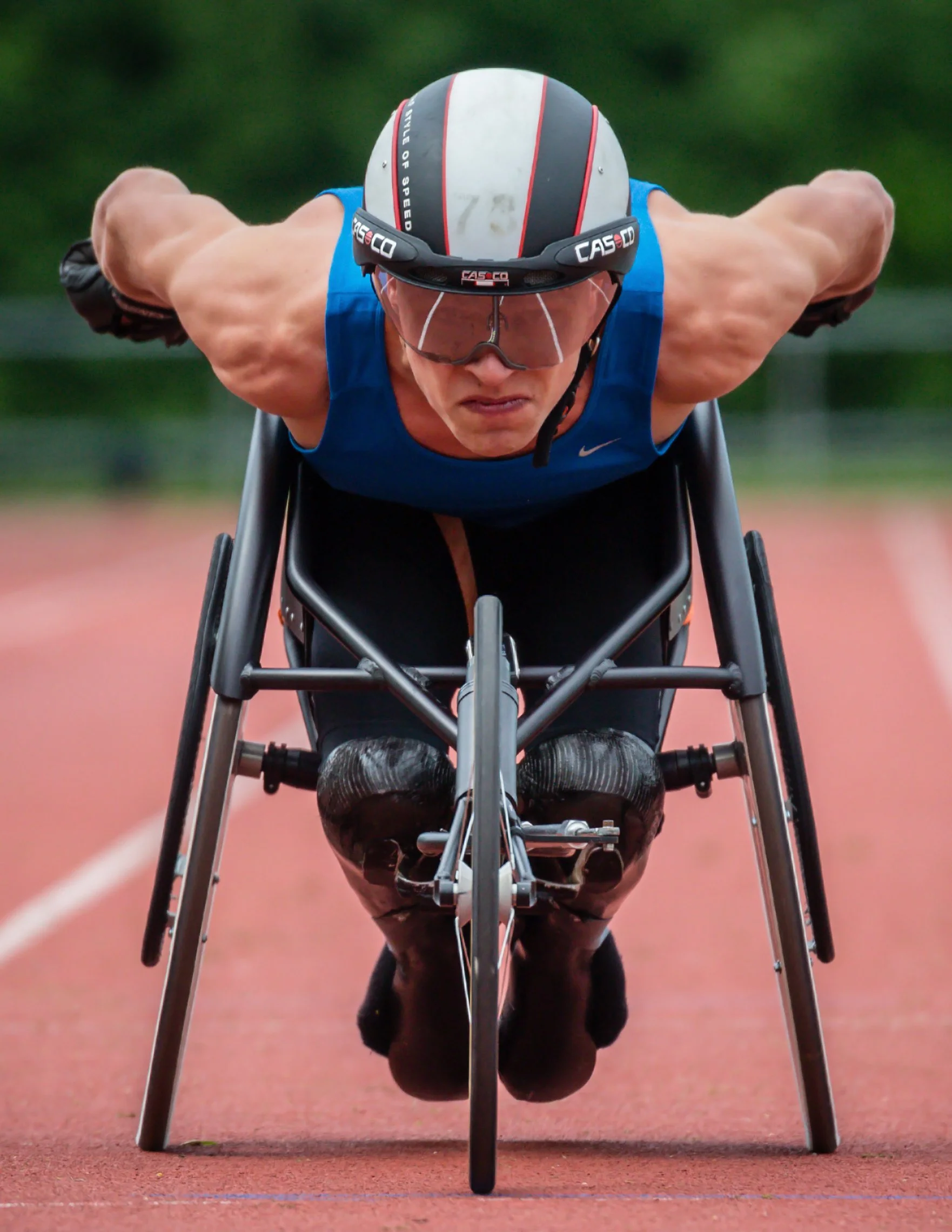 A male athlete in a racing wheelchair, wearing a helmet and protective glasses, leaning forward during a sprint on an outdoor track with blurred green background.