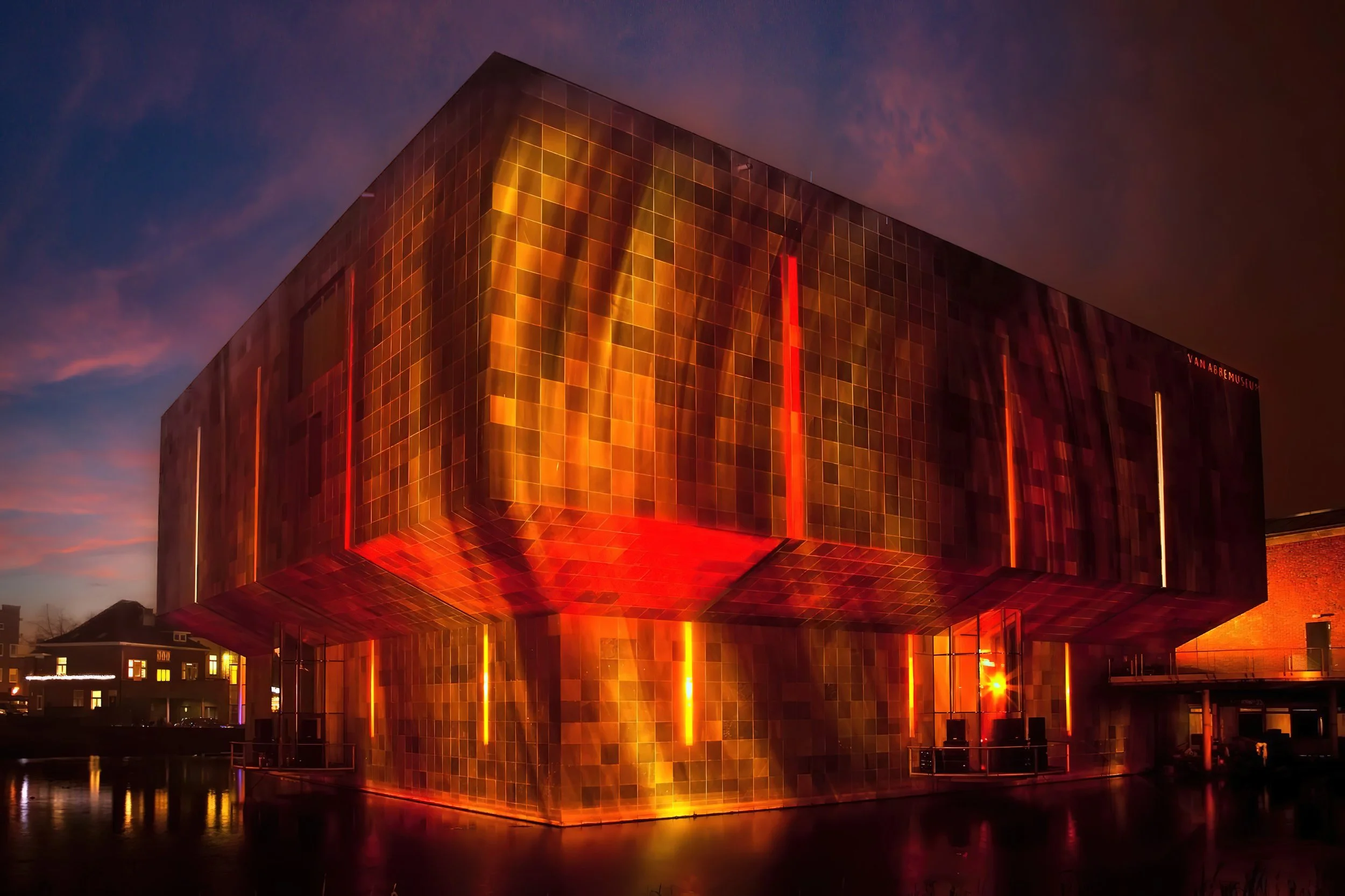 Night view of the Van Abbemuseum, a modern building with a reflective, golden-orange tiled exterior, lit from within, located beside a body of water.