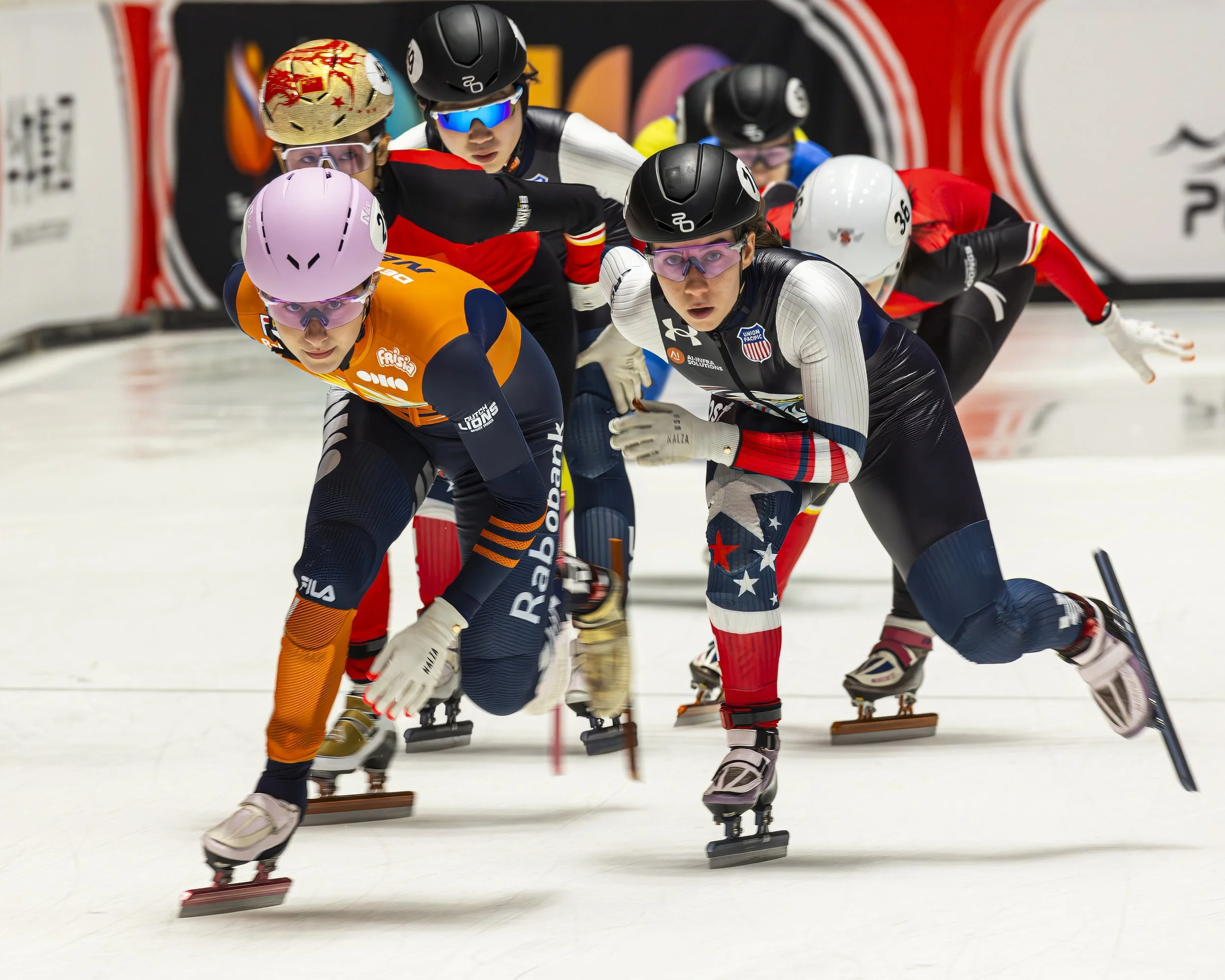 Speed skaters in a competitive race on an indoor ice rink, leaning forward in a tight formation, wearing colorful uniforms, helmets, and protective eyewear.