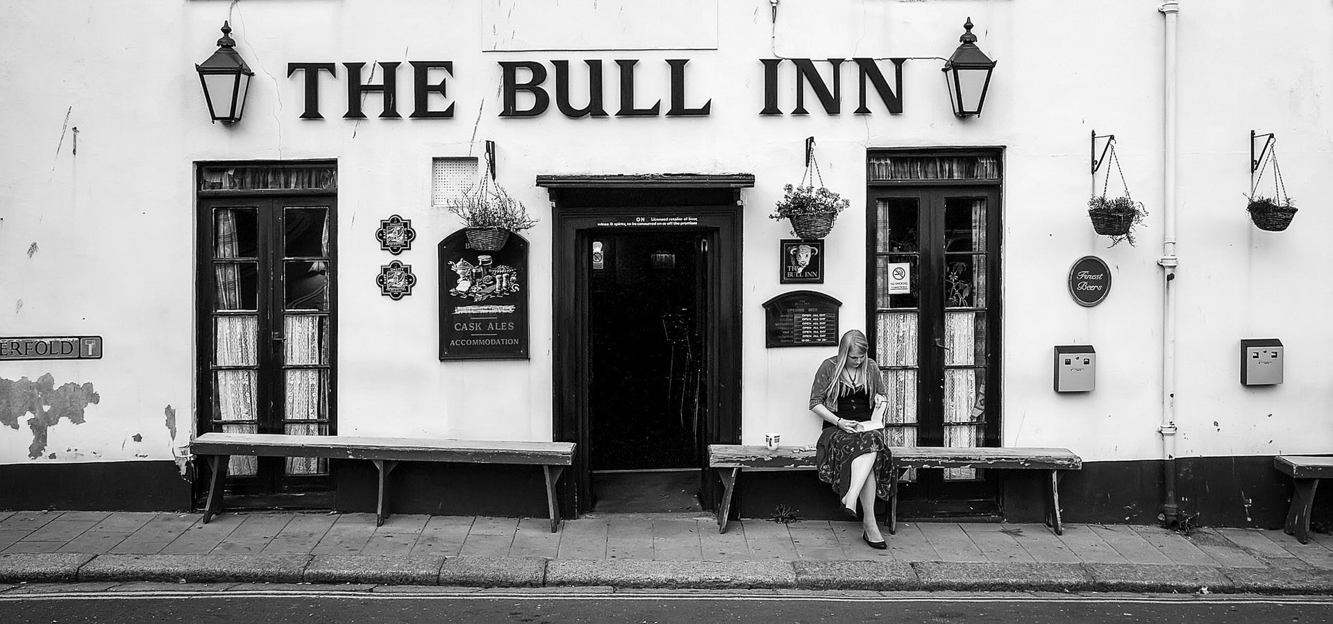 Black and white photo of an old pub called The Bull Inn. The pub's sign features a drawing of a bull. Two hanging lanterns are mounted on the wall beside the sign. There are windows with curtains, and small signs and decorations, including a sign off