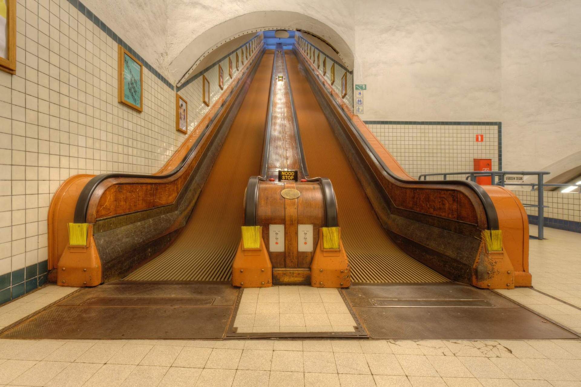 An old wooden escalator in a subway station, with steps that move upward and downward on parallel tracks, surrounded by tiled walls and floor. There are warning signs and safety barriers near the escalator.