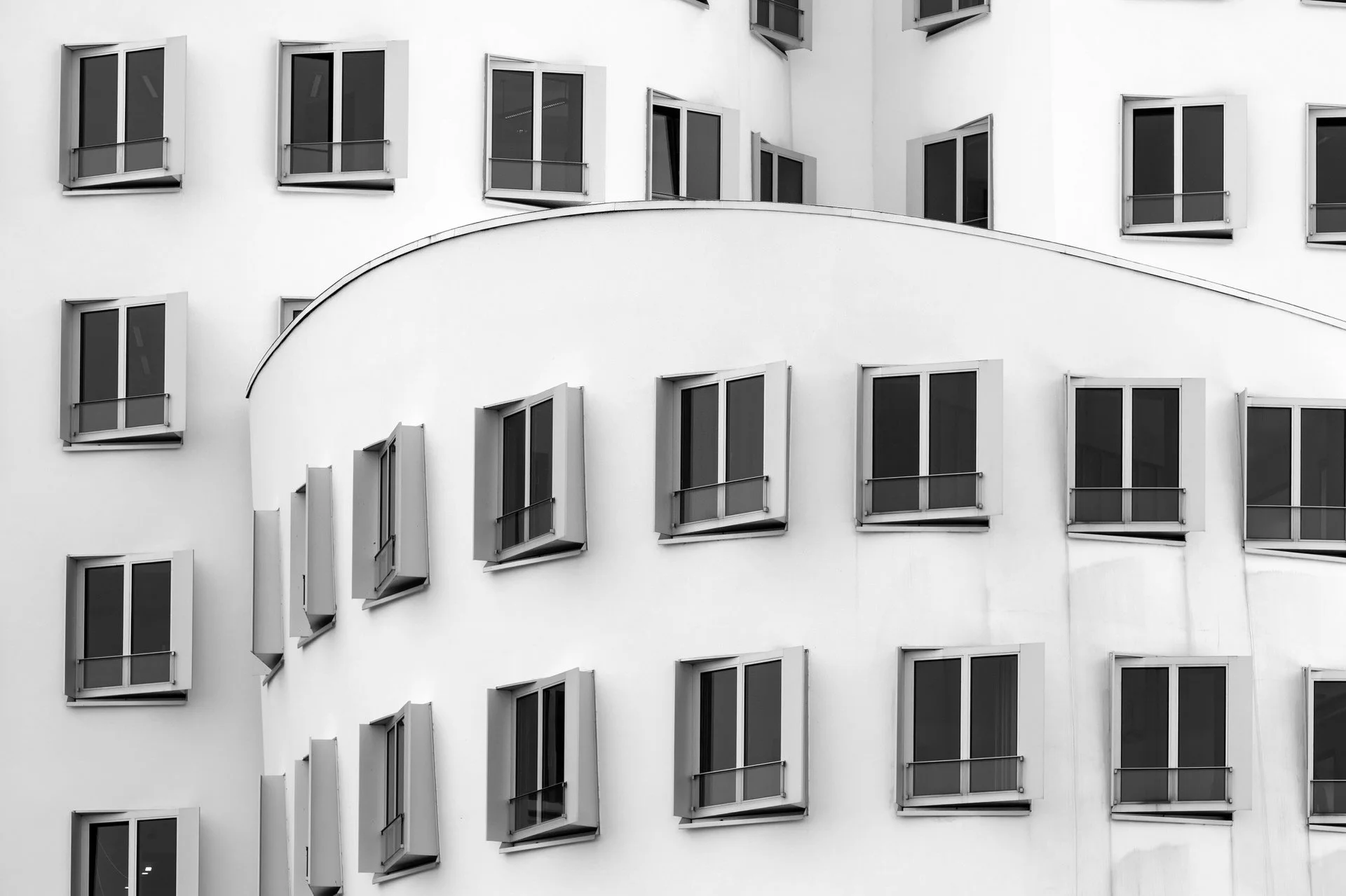 Black and white photograph of a modern building with multiple windows, some open and some closed, arranged in an irregular pattern on the white facade.