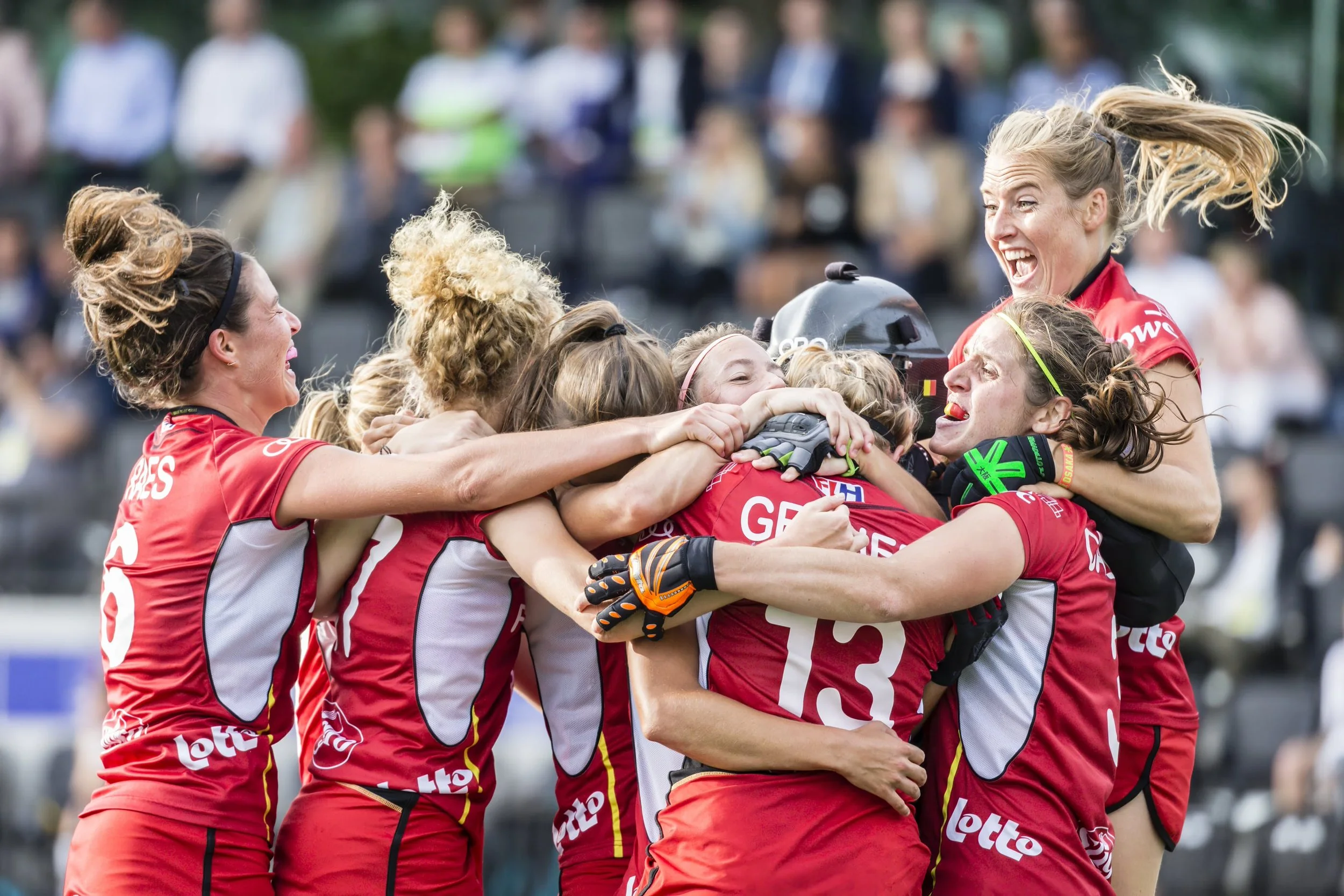A group of female athletes in red jerseys celebrating together in a huddle, with some lifting one woman in the air, outdoors with blurred spectators in the background.