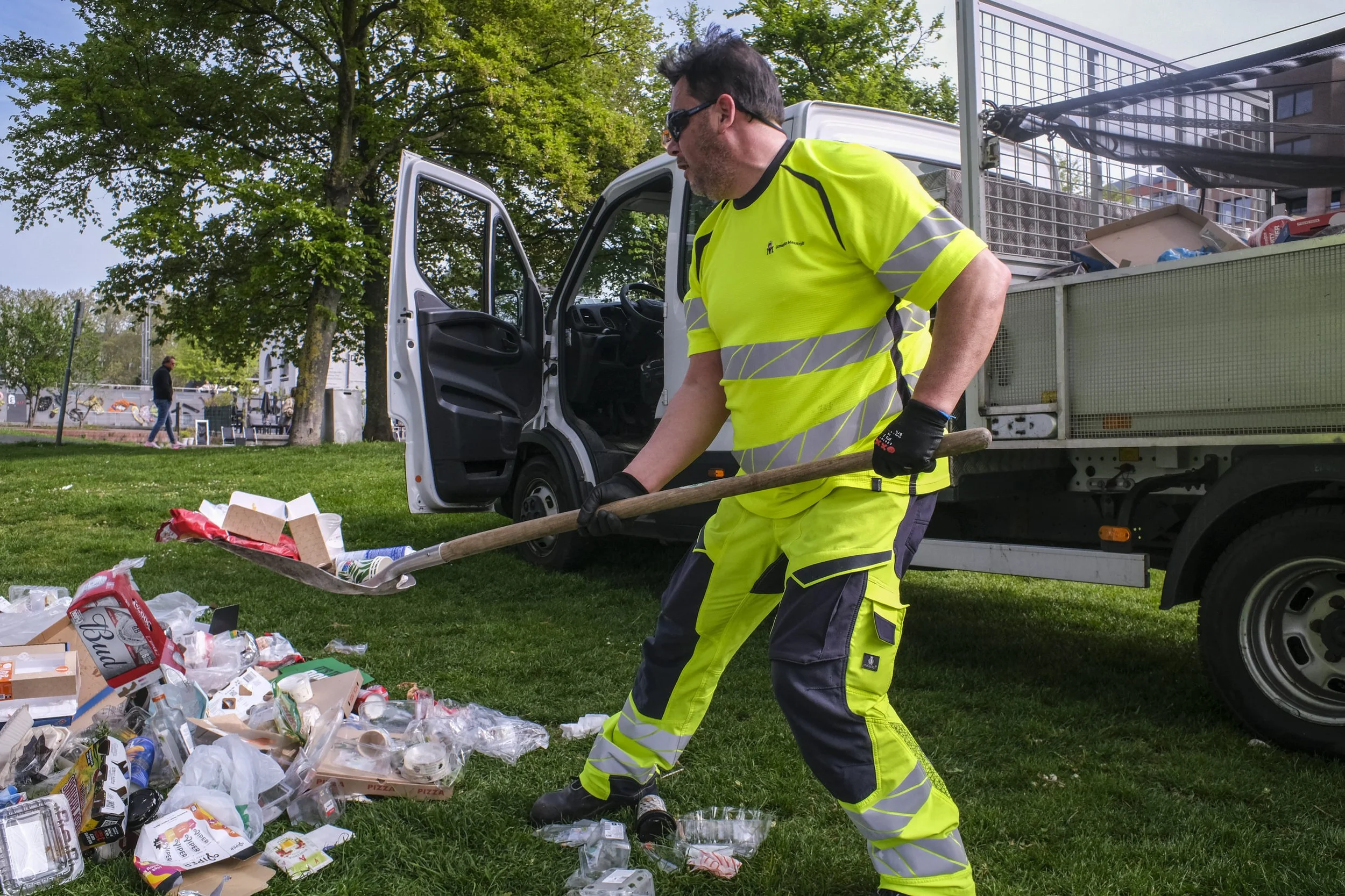 A man in high-visibility yellow and black safety clothing is cleaning up broken glass and trash from the ground near a data truck, with a park and a person walking in the background.