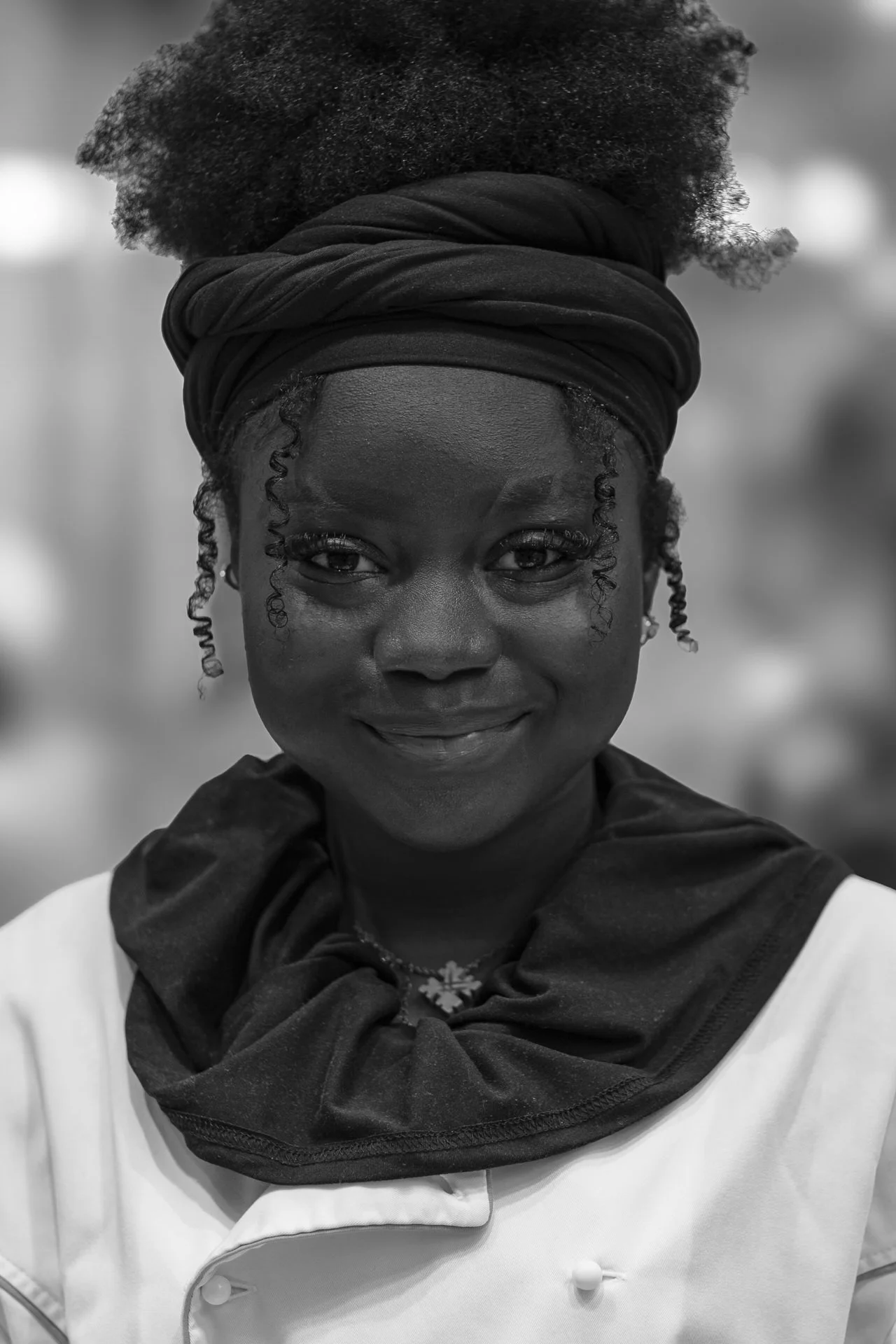 A woman with dark skin, curly hair wrapped in a headscarf, wearing a chef's uniform, smiling, and standing outdoors.