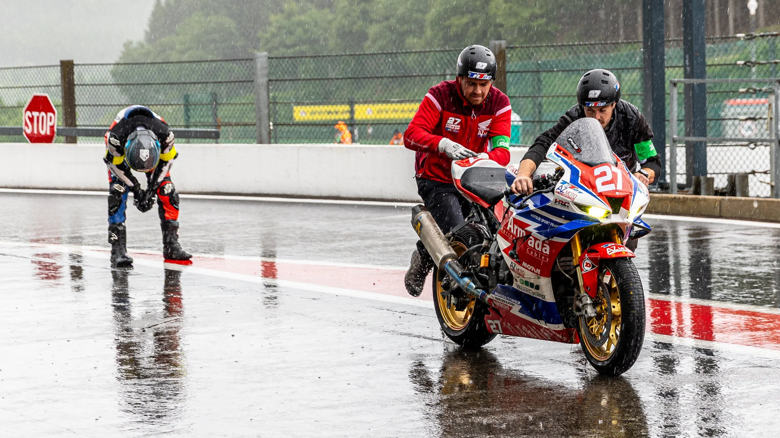 Race car and motorcyclist in the rain on a wet track, with a person in racing gear bending down in the background.