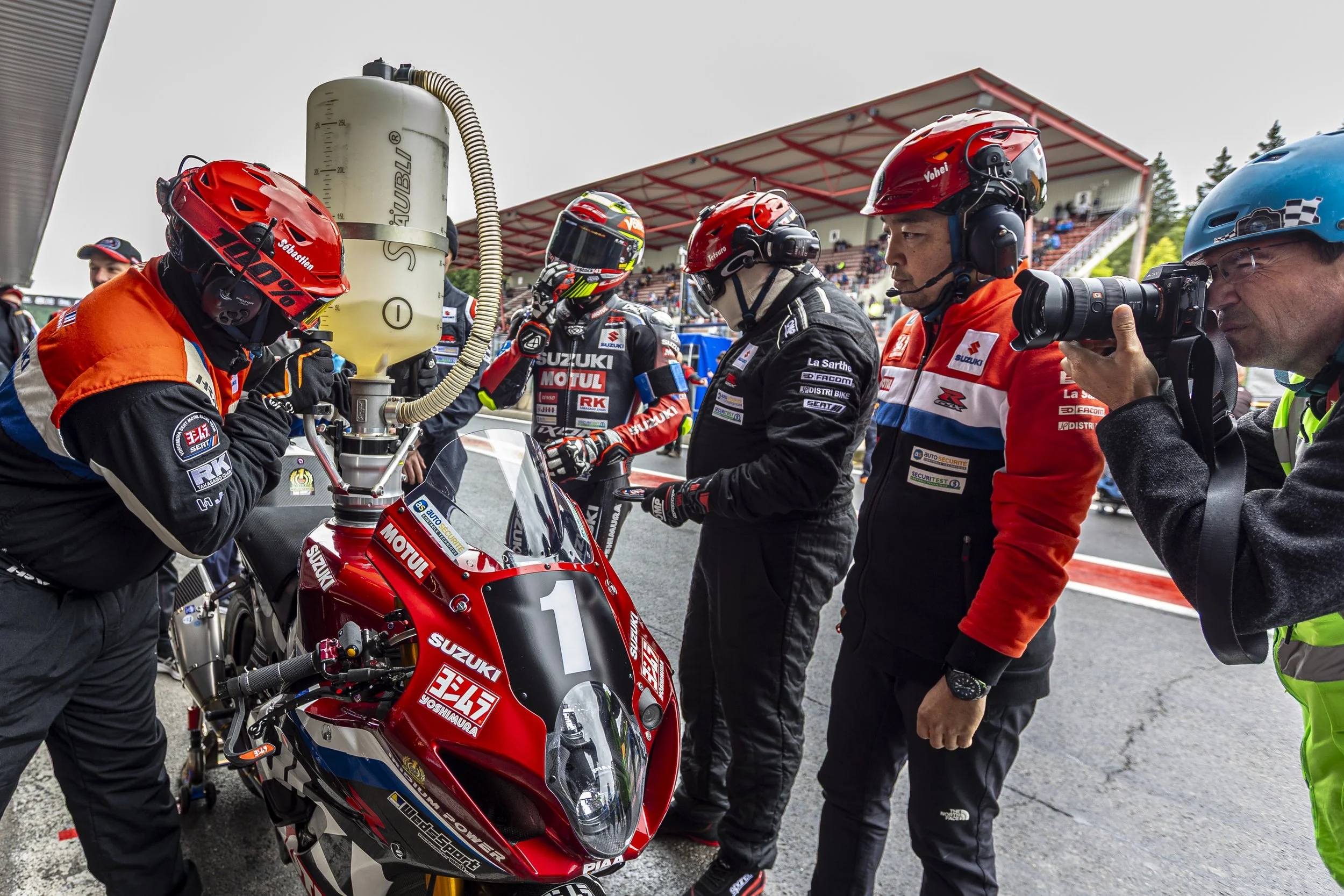 Race team members preparing a red Suzuki motorcycle with the number 1 logo before a race, with a rider wearing a helmet and race suit, in front of a grandstand at a racing event.
