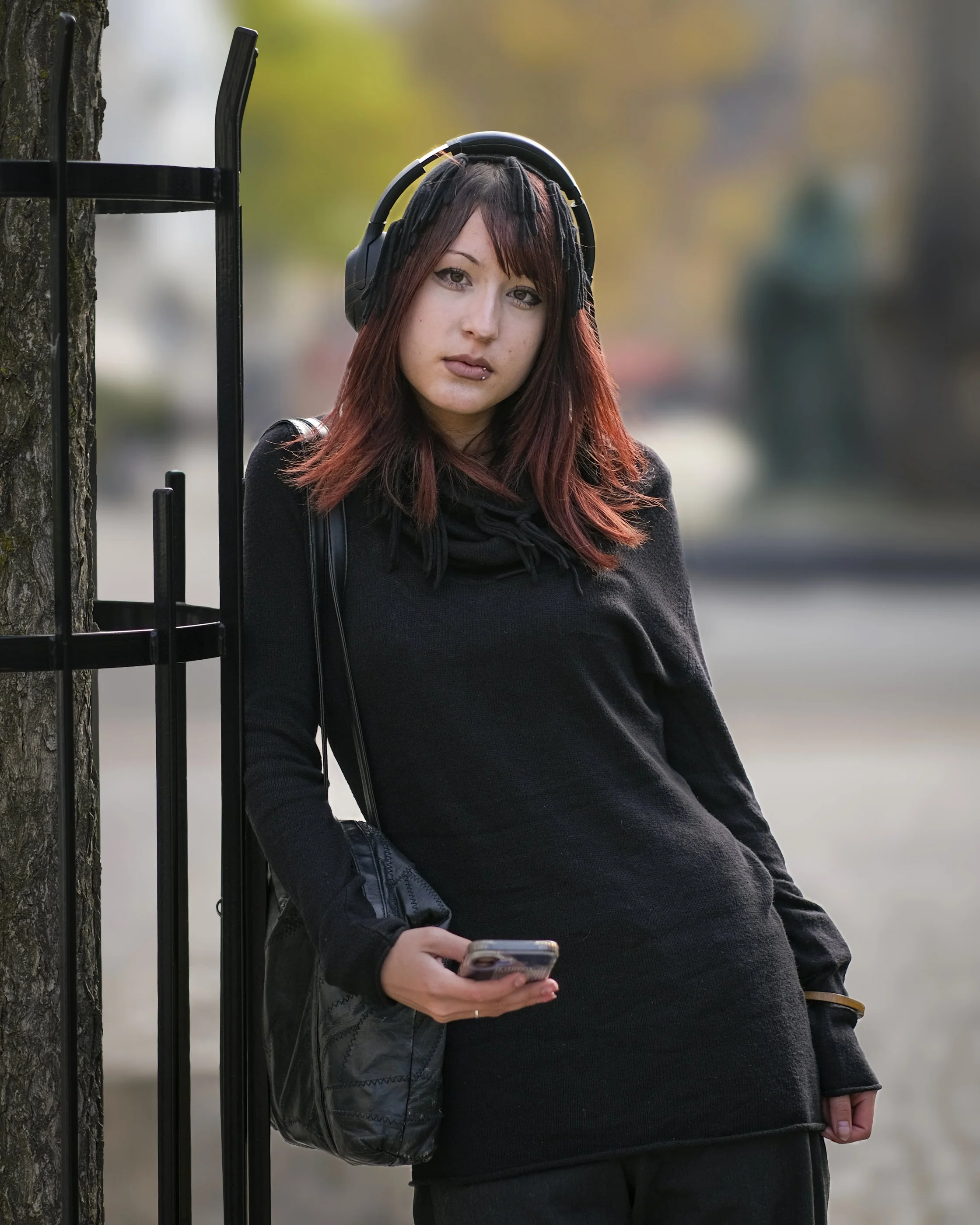 Young woman with red and black dreadlocks wearing headphones and black outfit, holding a smartphone, leaning against a tree with a metal fence in the foreground, blurred outdoor background.