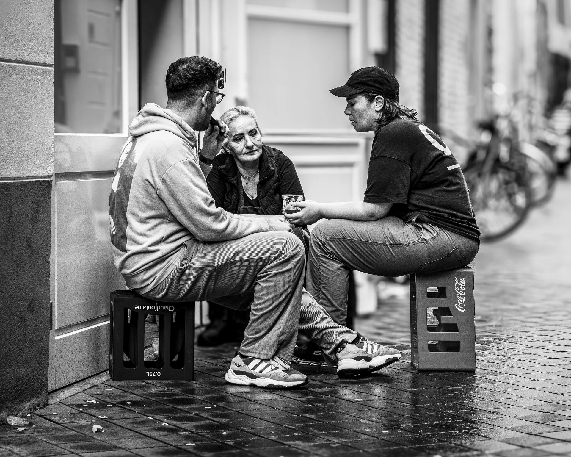 Three people sitting on crates on a city street, engaged in conversation. The scene is in black and white.