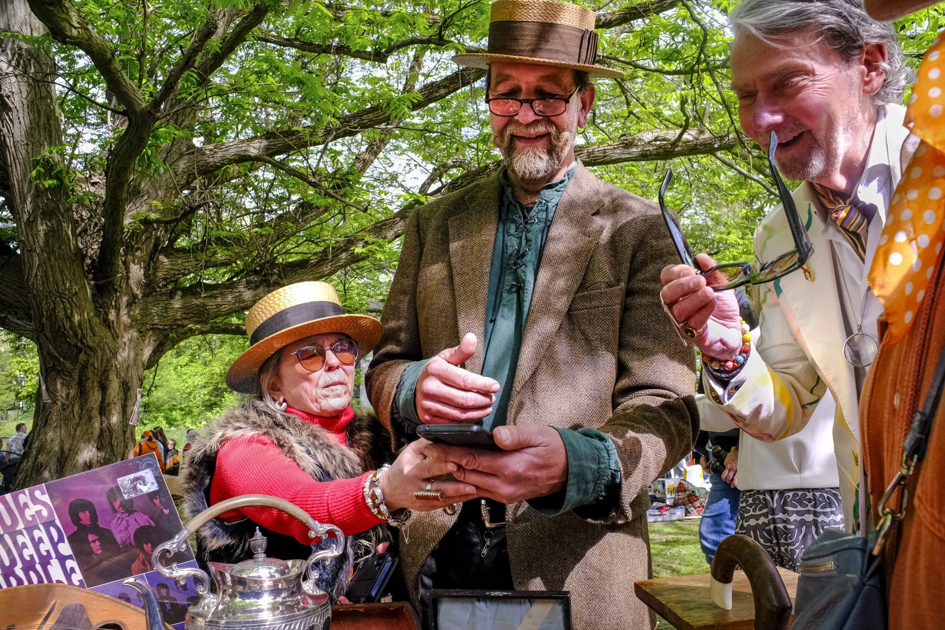 A group of people at an outdoor event looking at a smartphone, surrounded by trees and greenery. One man has gray hair, glasses, and a striped tie; another woman wears a hat and sunglasses; a third man has a tall hat and beard.