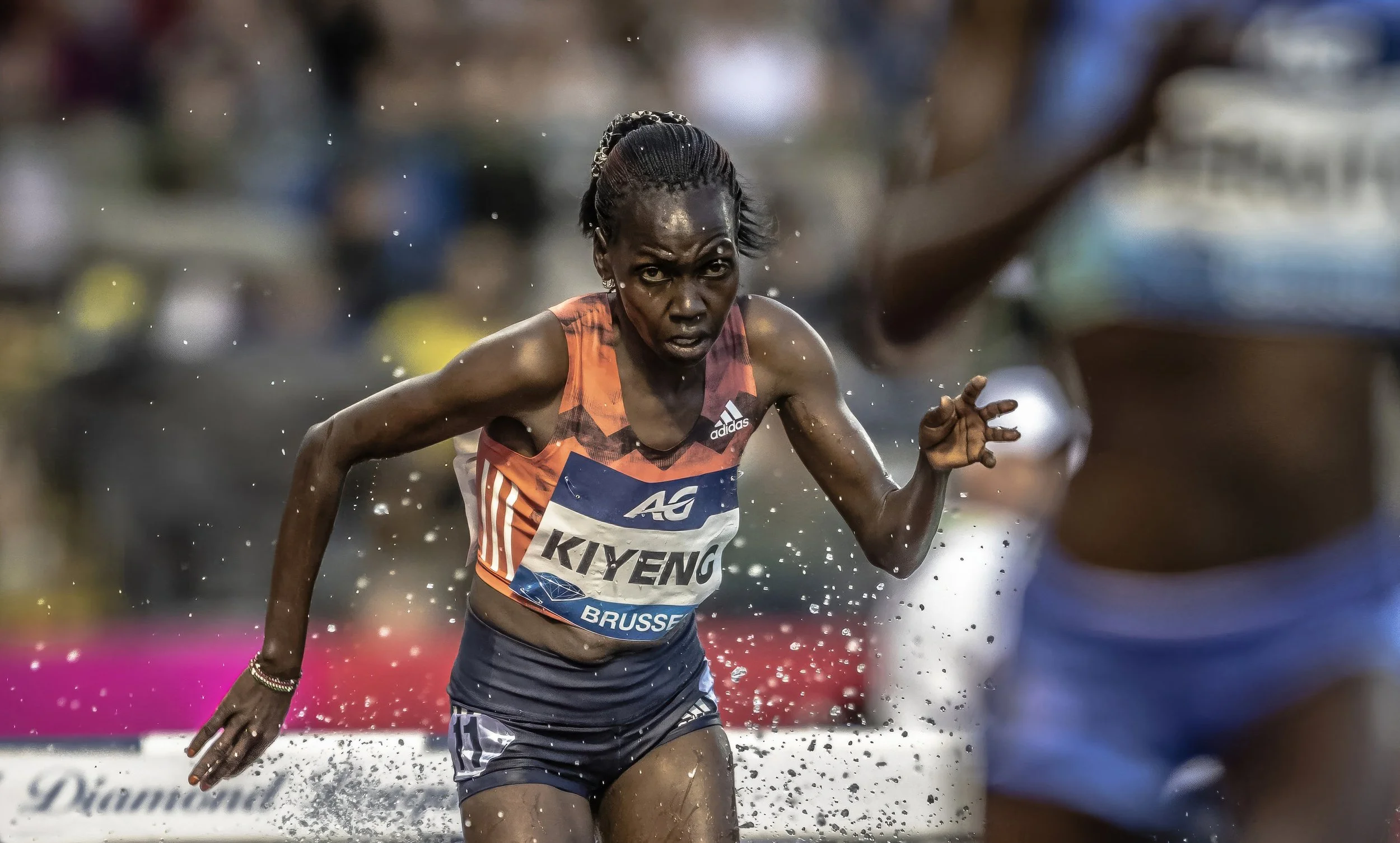 An athletic woman running through water during a race, wearing a red and black sports top with the name Kiyeng and the word Brussels, with water splashing around her and a blurred crowd in the background.