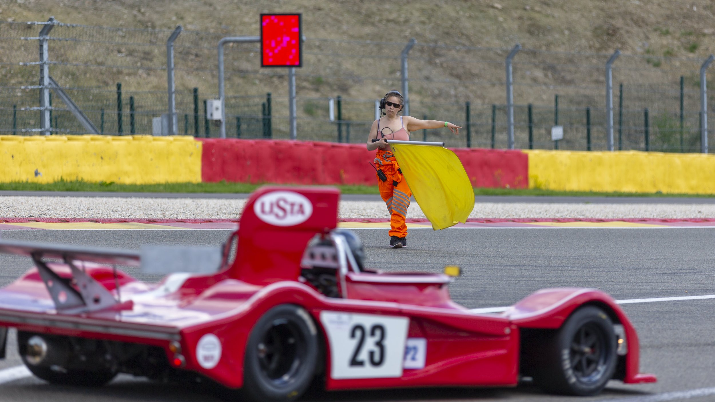 A person wearing sunglasses, a headset, and orange racing pants is standing on a race track, pointing towards a red Ferrari race car in the foreground. The track has red and yellow barriers and a digital red warning sign on a pole in the background.
