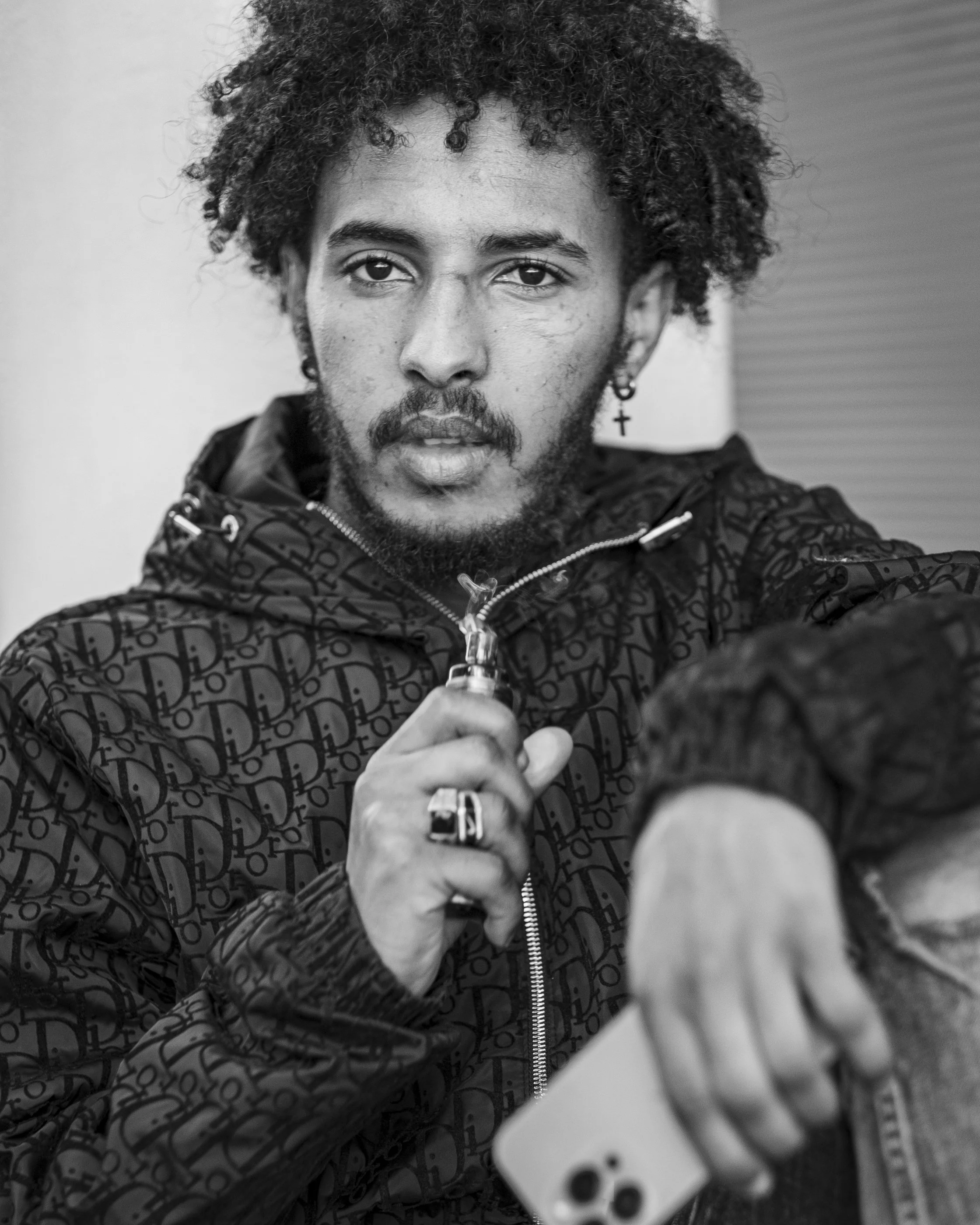 Black and white photo of a young man with curly hair and a cross earring, taking a selfie with a confident expression. He is wearing a patterned jacket and holding a smartphone in his hand.