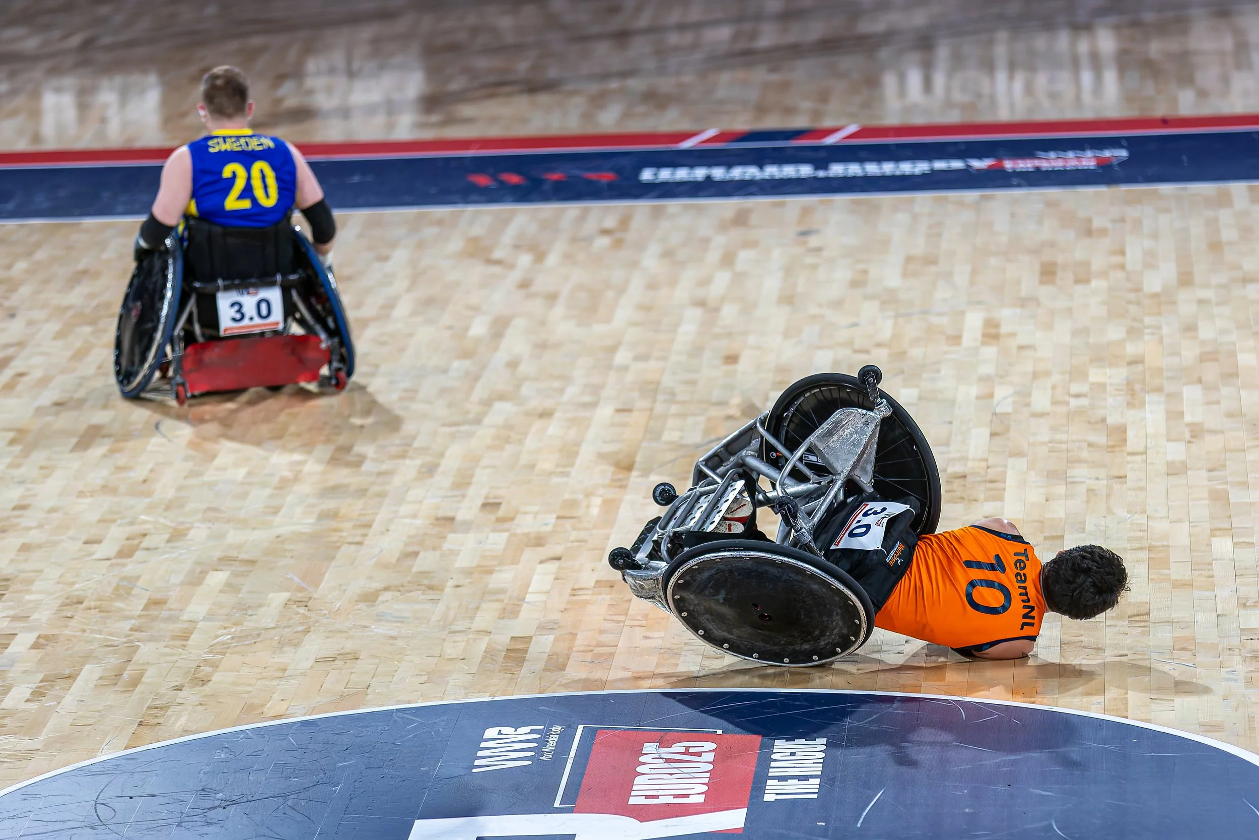 A wheelchair user falling onto a basketball court during a game, with their wheelchair overturned nearby. Two other players are visible on the court.