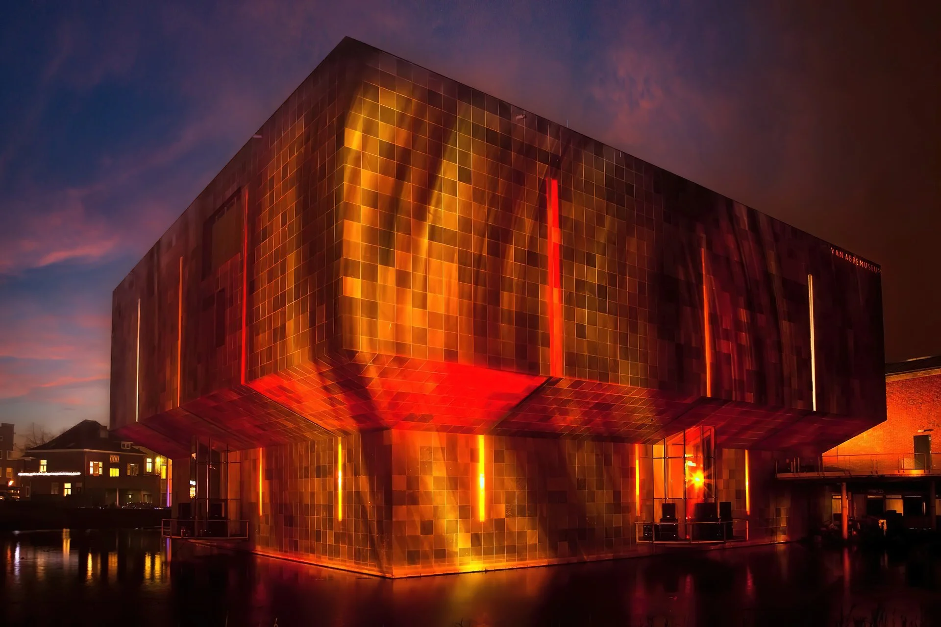 The Van Abbe Museum, a large modern building with a glowing orange and red metallic facade, illuminated at sunset in the night sky.
