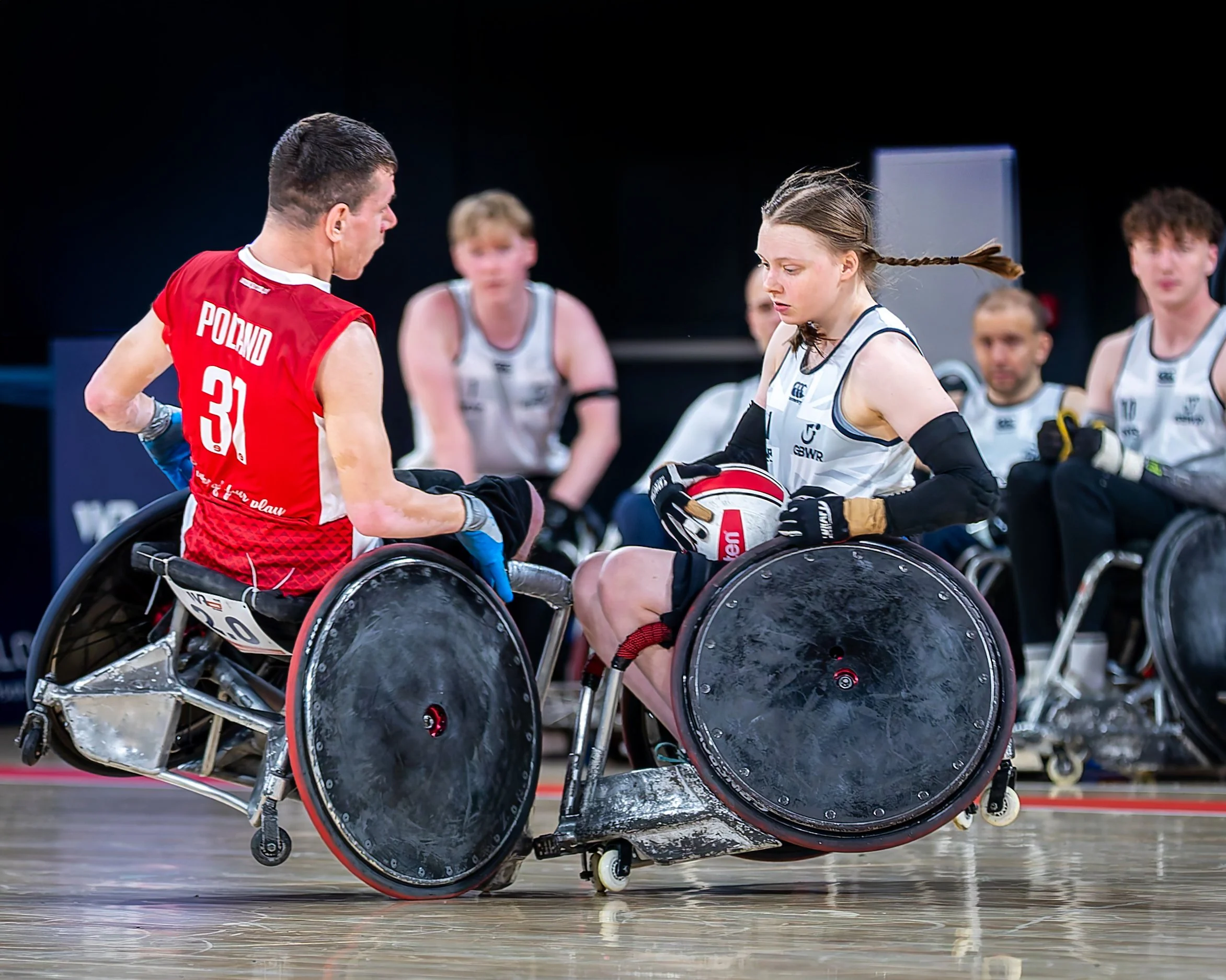 A female athlete in a wheelchair holding a red and white volleyball, surrounded by teammates and coaches during a game or practice, indoors with a dark background.
