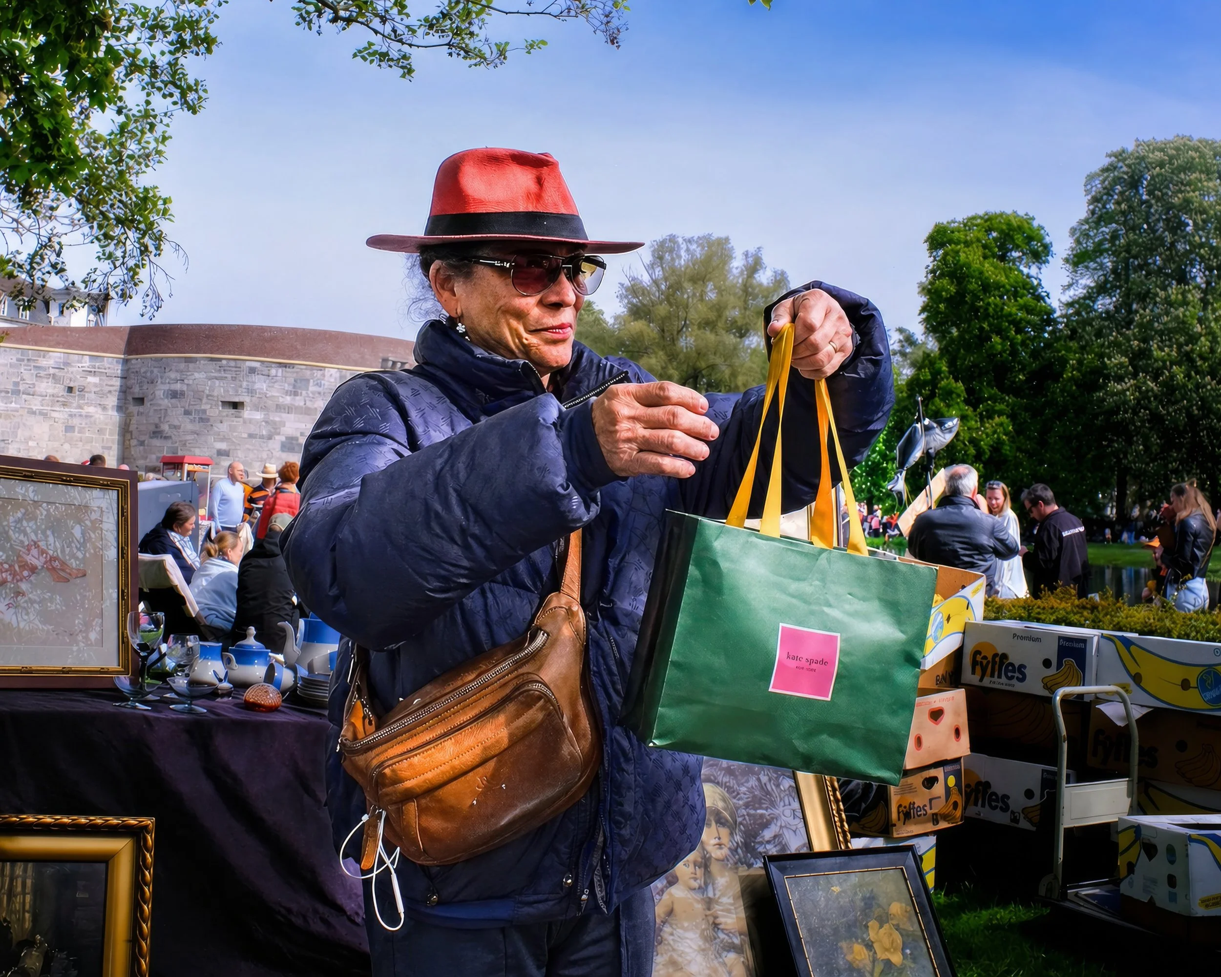 A woman in a red fedora and sunglasses shopping at an outdoor market, holding yellow shopping bags, with other people and market stalls visible in the background.
