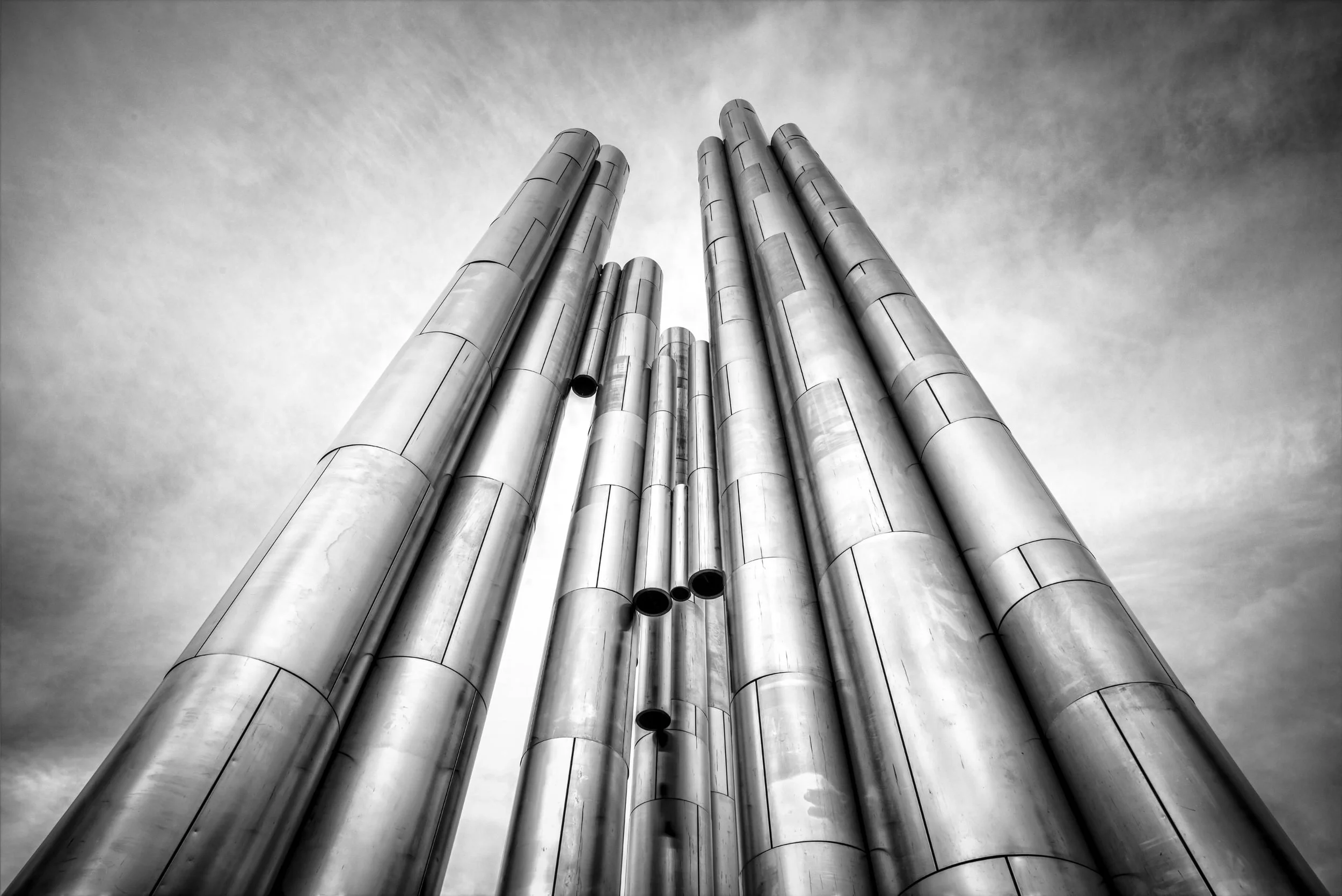 Black and white photo of large cylindrical metal pipes extending upward towards the sky, with some pipes open at the ends.