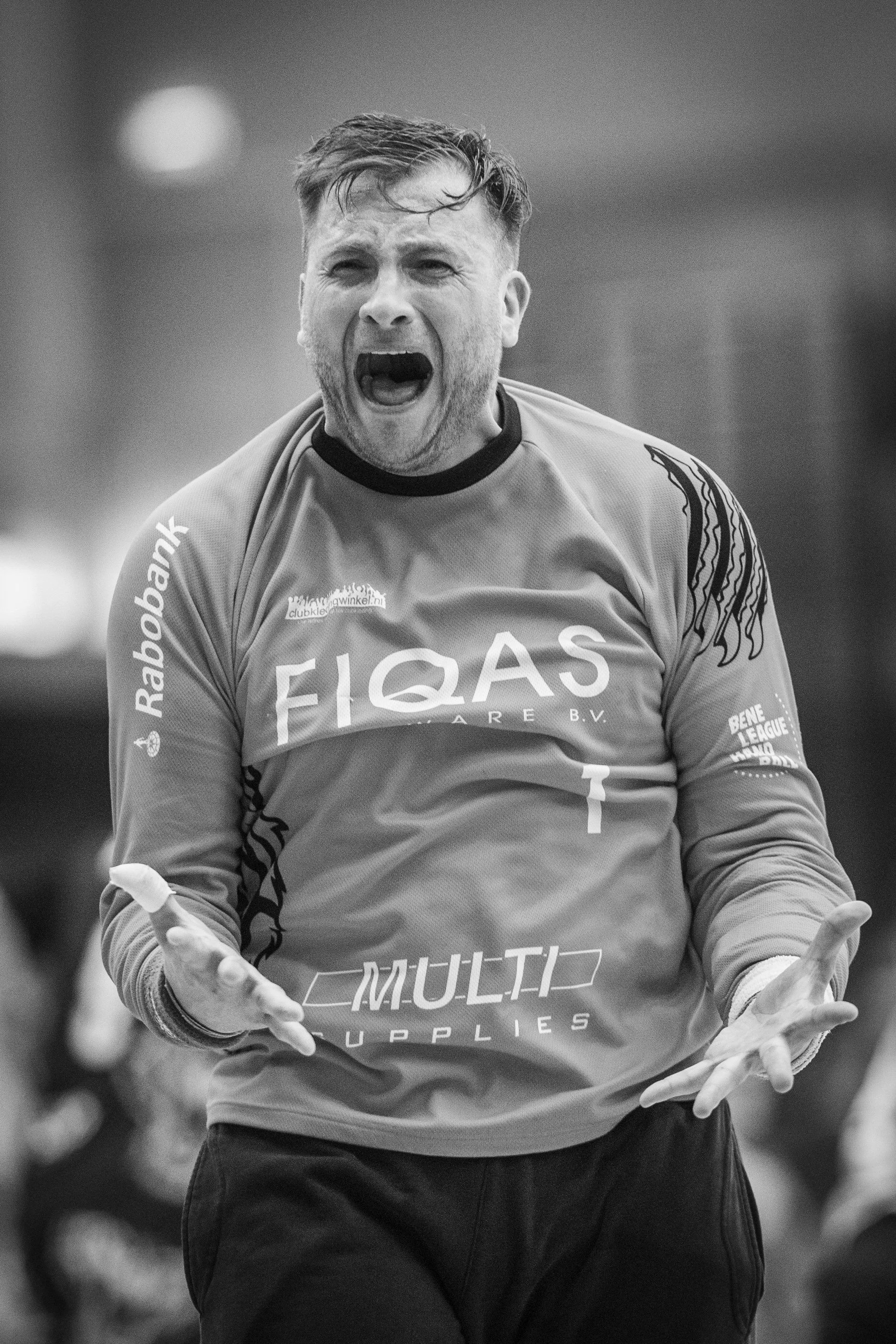 A black and white photo of a man with short hair, wearing a sports jersey, expressing a strong emotion with his mouth open and hands gesturing.
