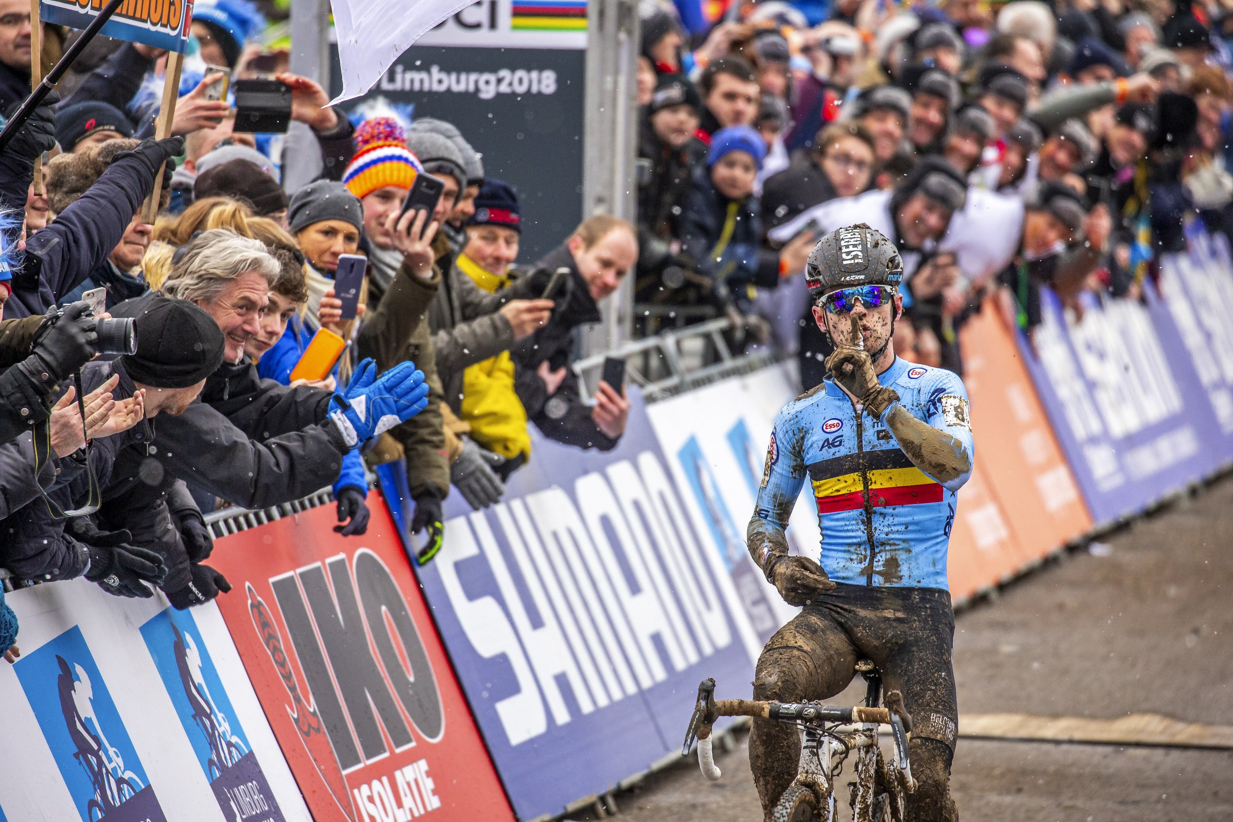 Cyclist covered in mud crossing a finish line with cheering spectators behind a barrier.