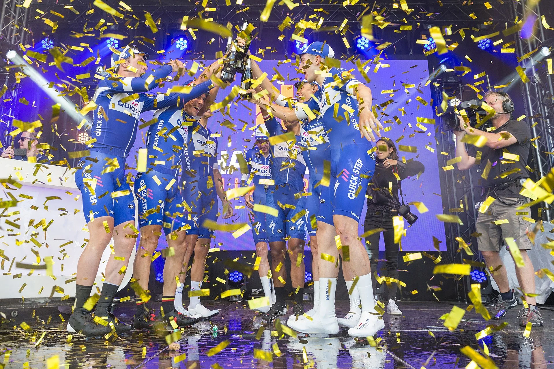 A group of cyclists in team uniforms celebrating a victory on stage, surrounded by falling yellow confetti, with a cameraman capturing the moment.