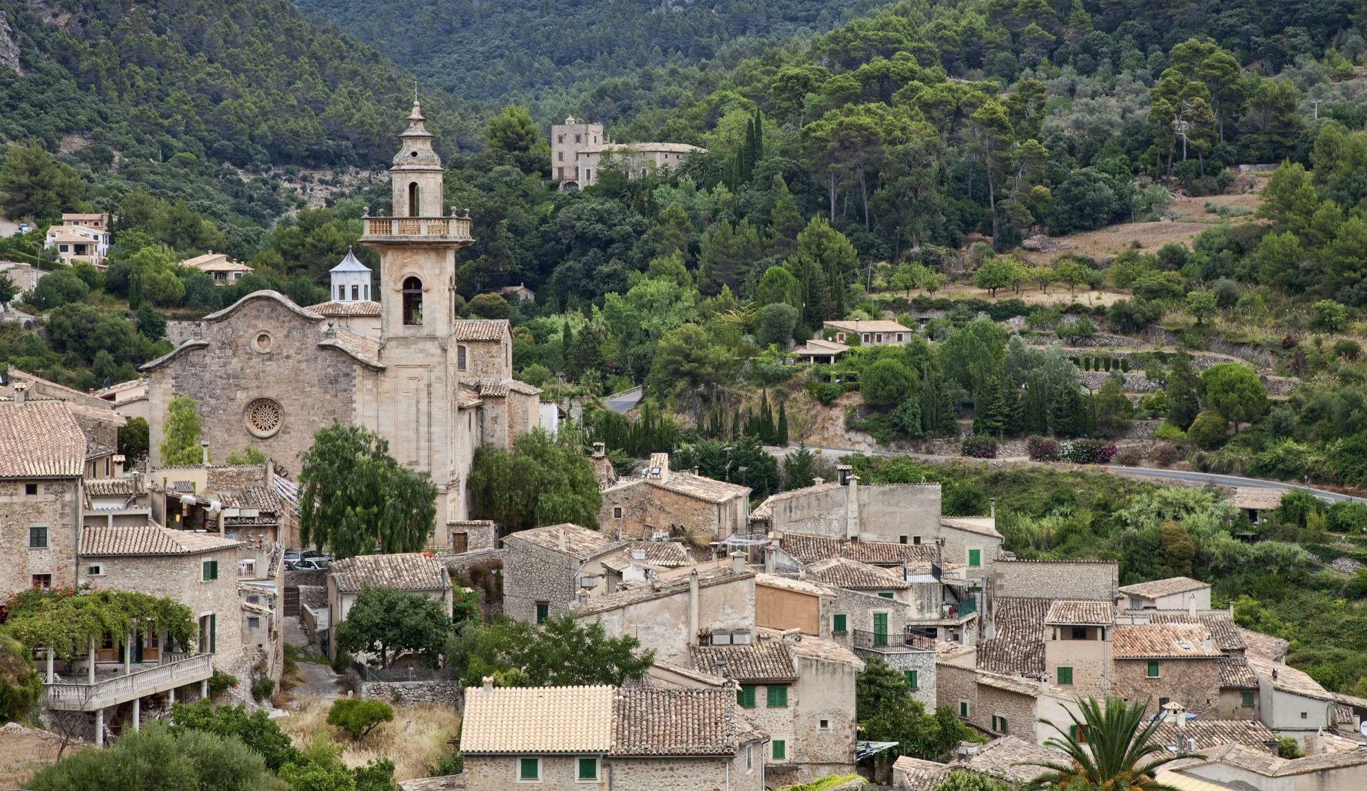 A hillside village with stone buildings, a church with a bell tower, and lush green trees and hills in the background.