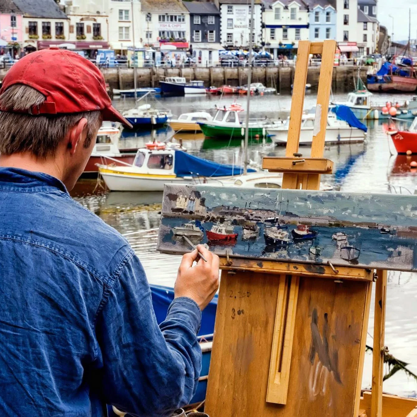 An artist in a red cap and blue denim shirt painting a harbor scene with boats, using an easel and canvas, with a coastal town in the background.