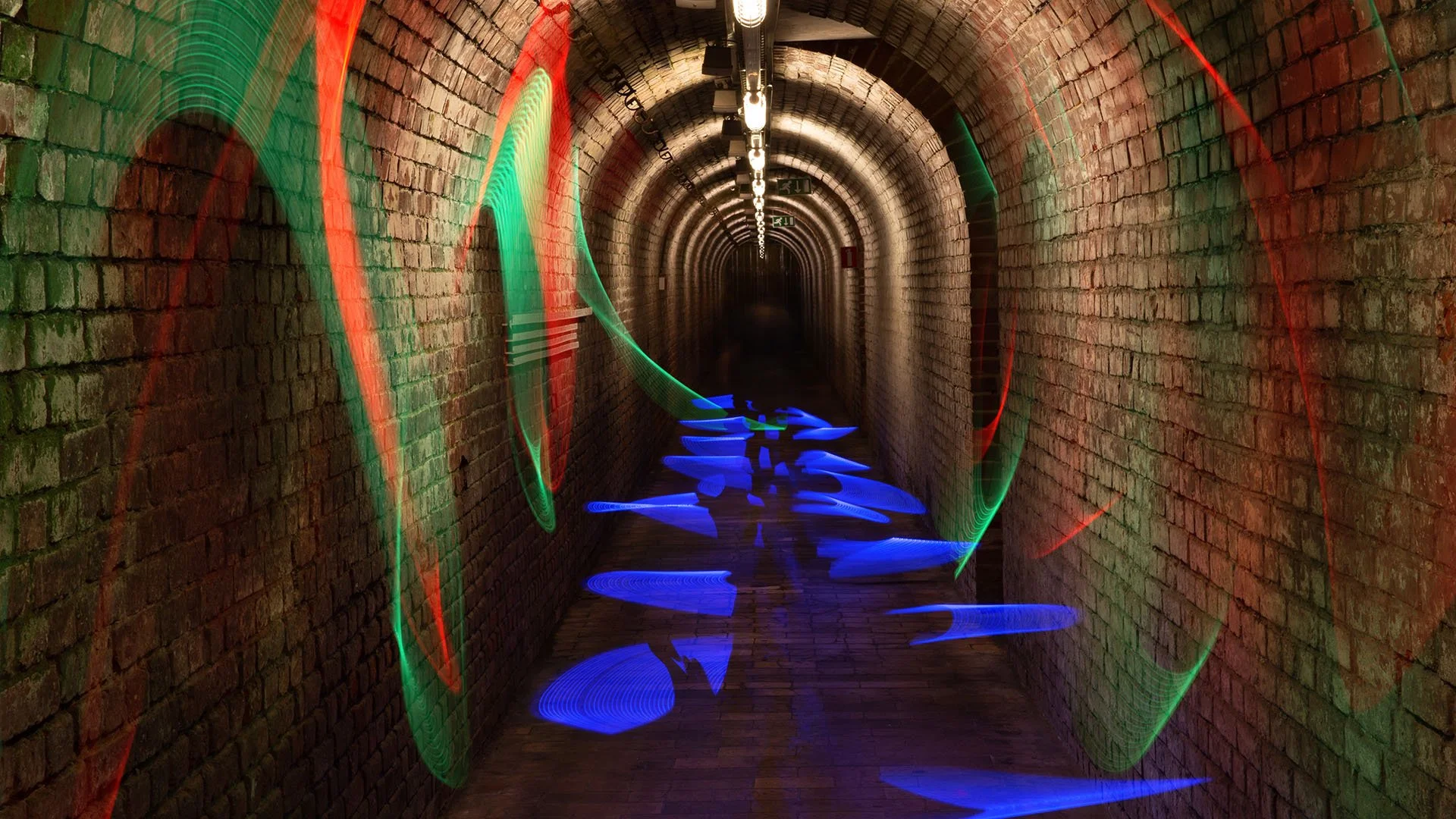 A long, narrow brick tunnel with colorful light streaks, including red, green, and blue, creating a light trail effect throughout the scene.