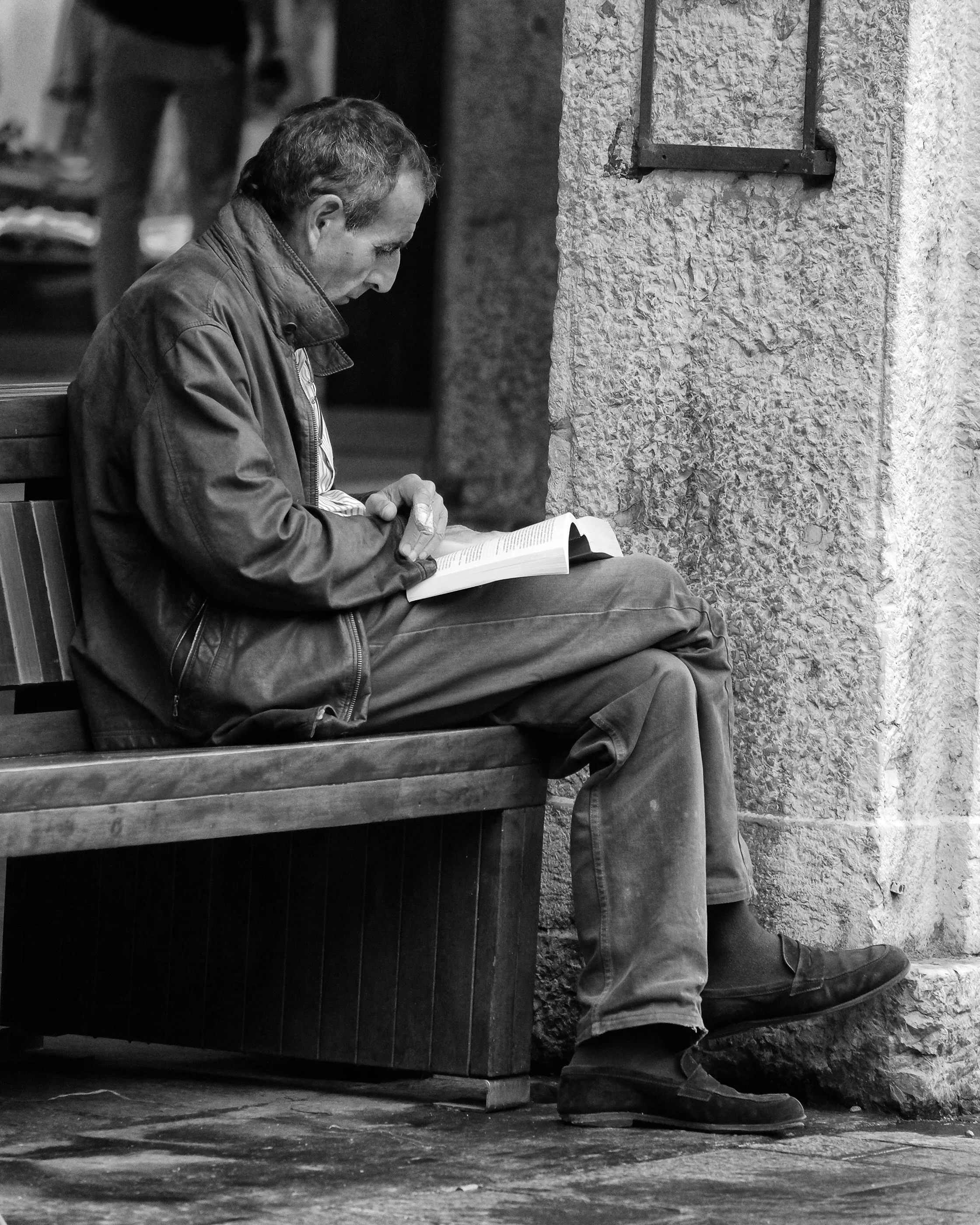 A man sitting on a wooden bench, reading a book, outside near a textured wall, wearing a jacket and boots, black and white photo.