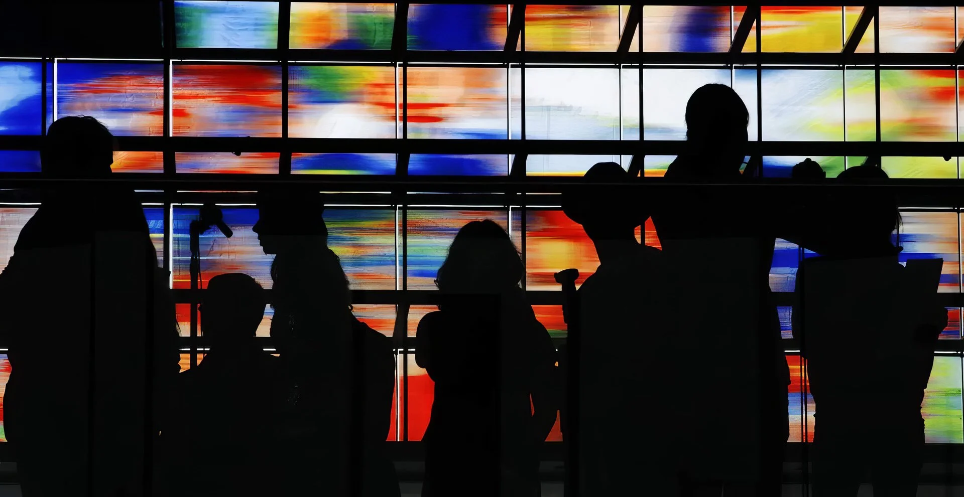 Silhouettes of people waiting at an airport terminal in front of a large, colorful abstract digital display screen.