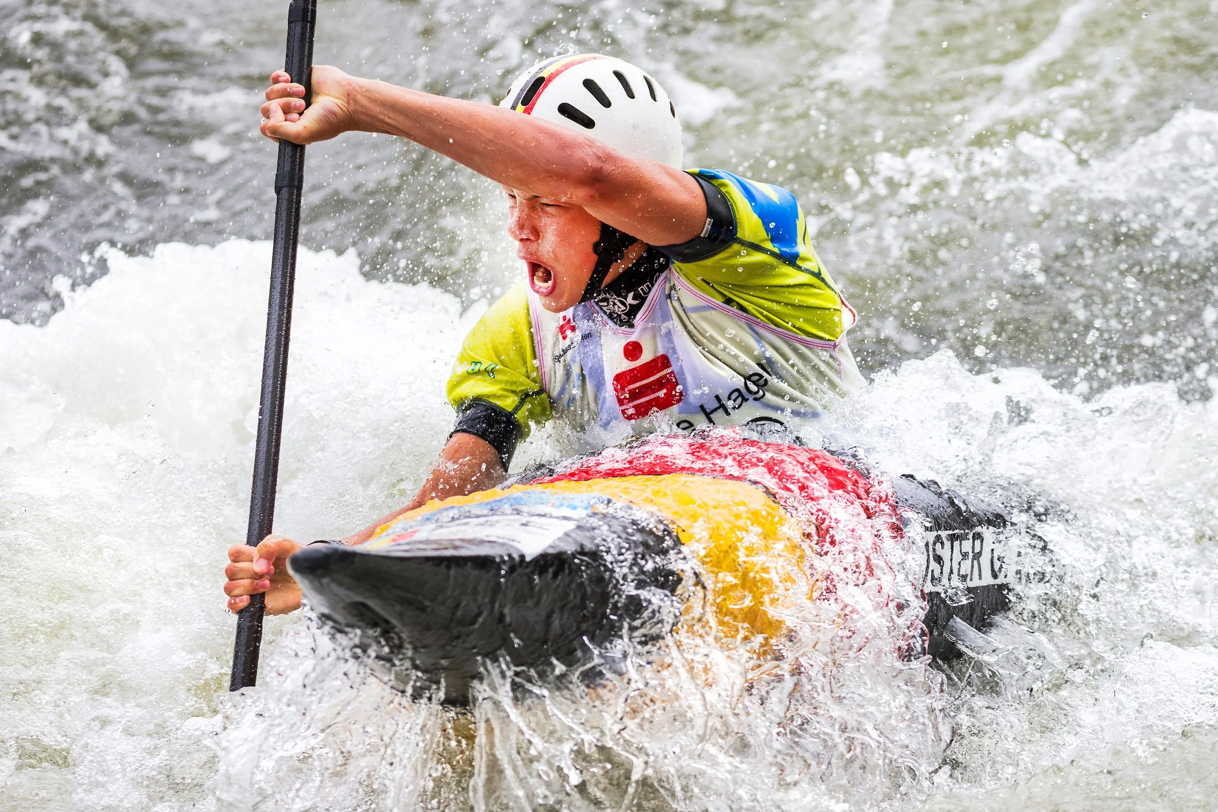 A person wearing a white helmet and a colorful life jacket is kayaking and appears to be struggling or falling into the water, with water splashing around them.