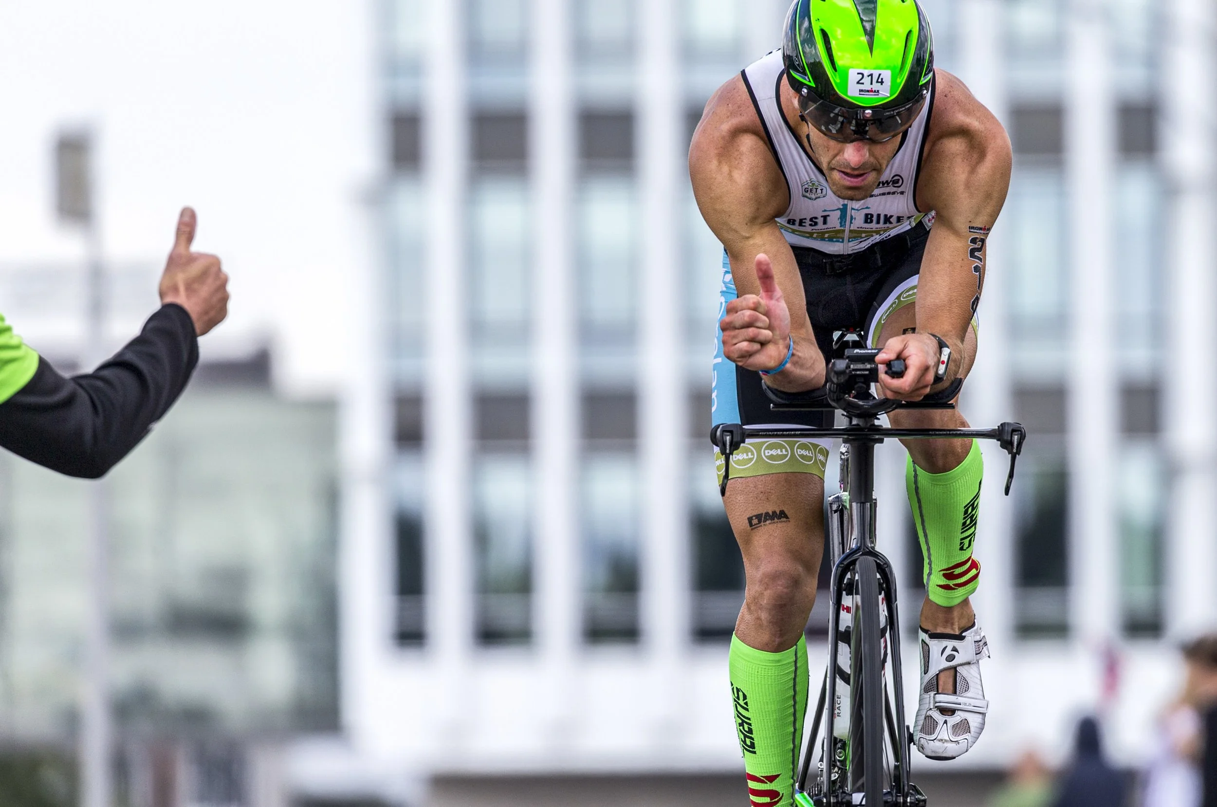 Male triathlete in neon green and black helmet, sunglasses, and athletic gear riding a bike during a race. He is giving a thumbs up, with a person in black and green near him. Urban background with blurred buildings.