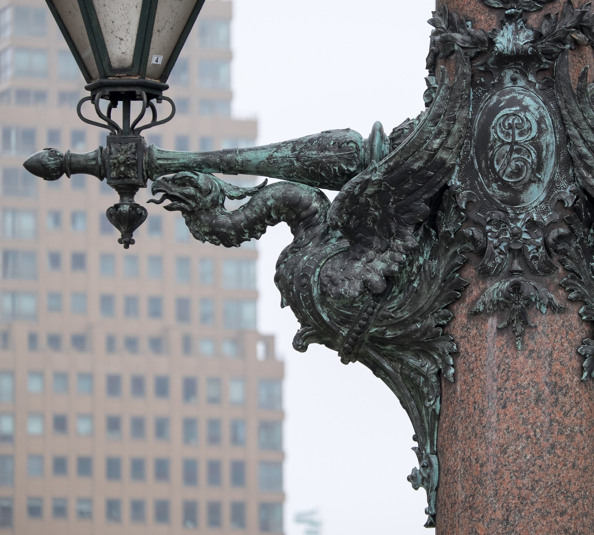 Close-up of an ornate outdoor lamp post with a decorative crest and lion head details, mounted on a reddish stone base, with modern office buildings in the background.