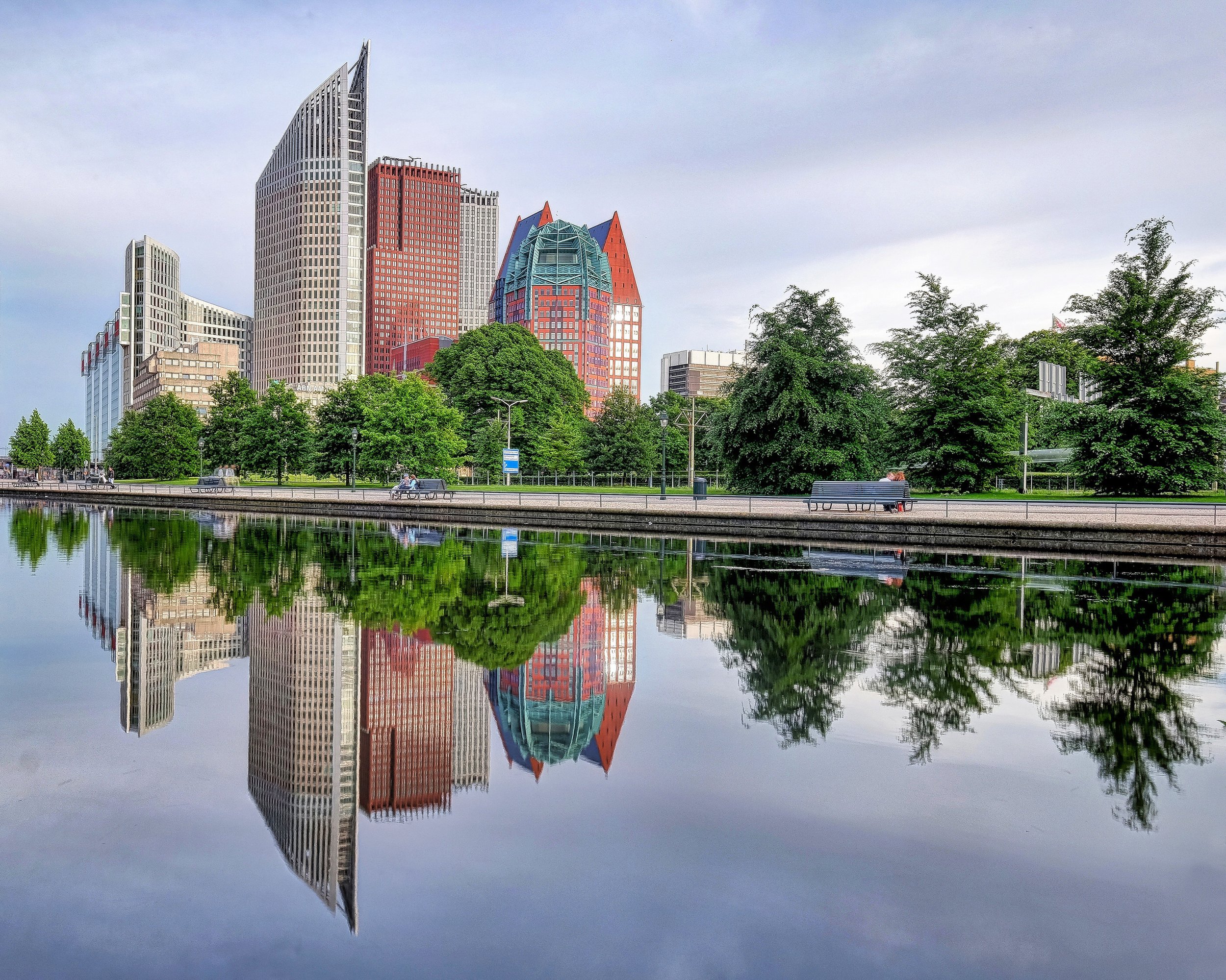 City skyline with modern skyscrapers reflected in a calm body of water, with green trees and park benches in the foreground.