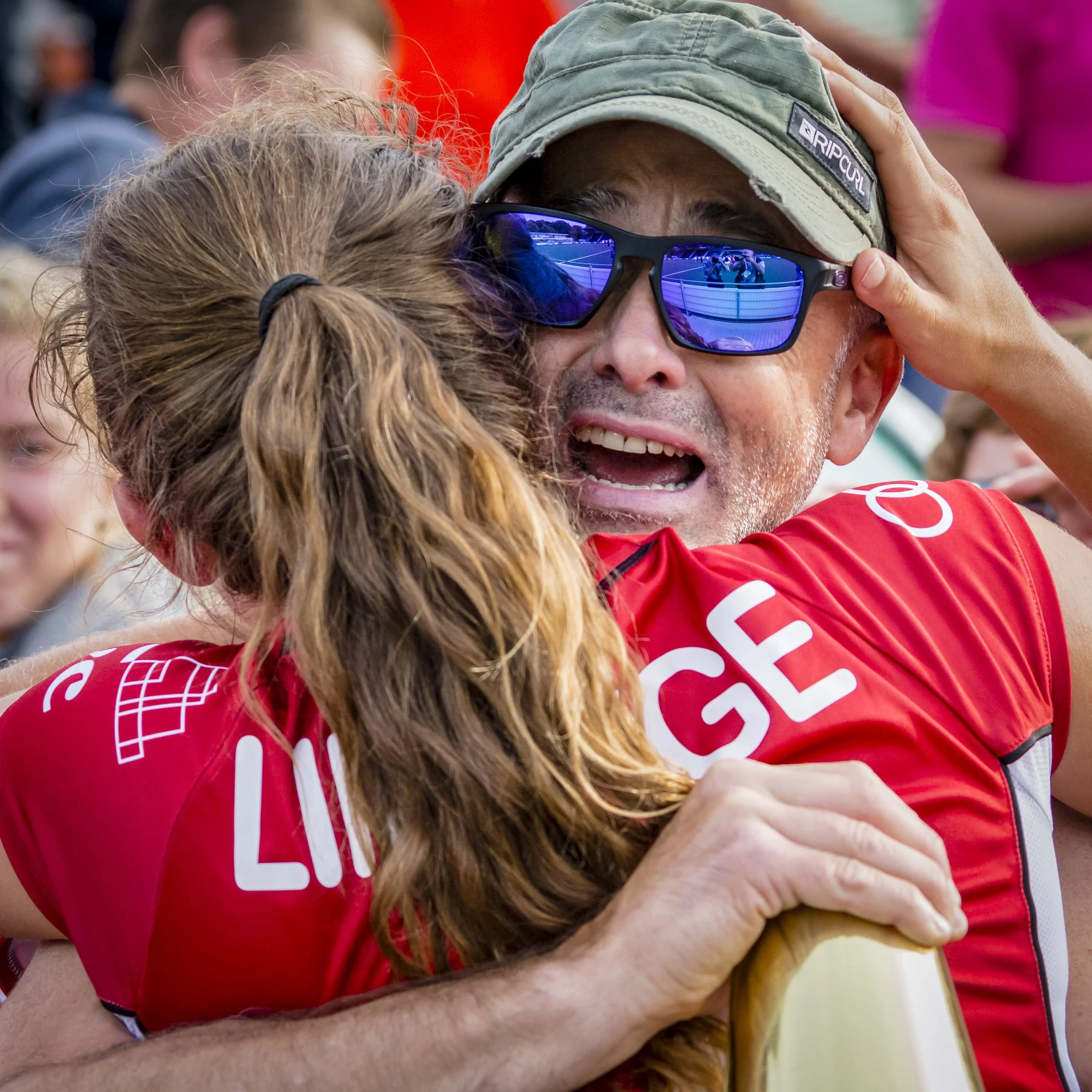 A person wearing sunglasses and a green cap hugging another person with long brown hair, with a blurred crowd in the background.