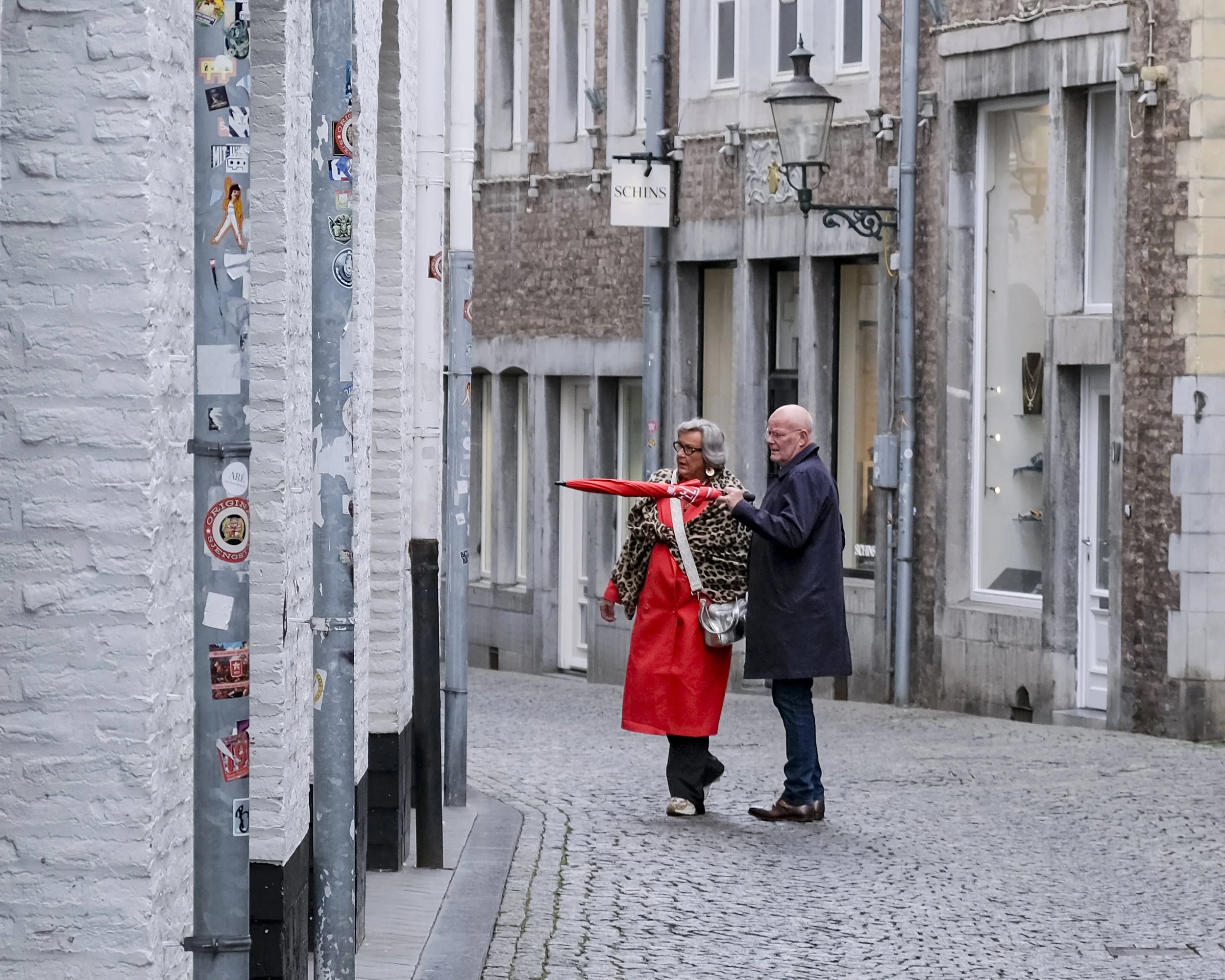 Two women are standing on a cobblestone street near a provencal wall with a sign that says "SCHINS." One woman is wearing a horse-print coat and red skirt, holding a red umbrella. The other woman is wearing a dark outfit and glasses. There are metal 