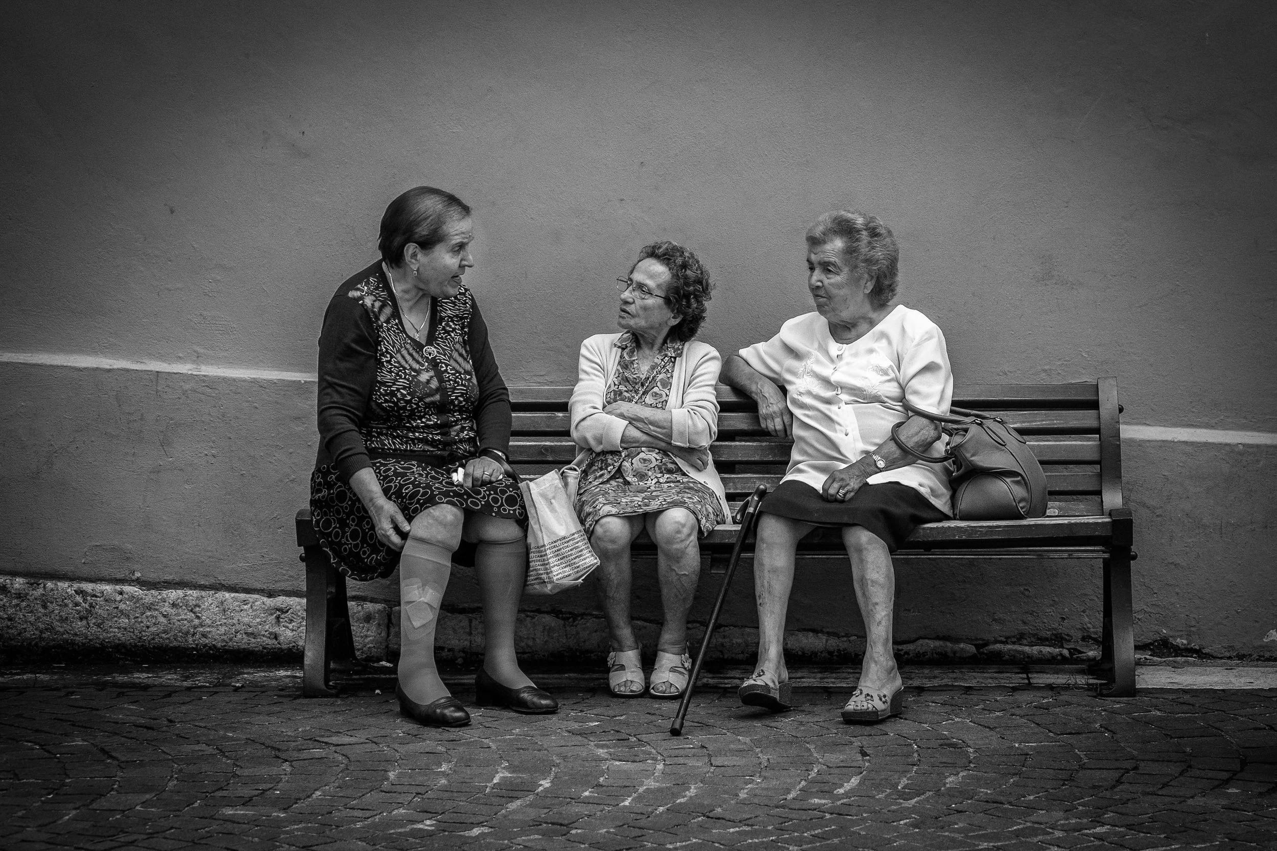 Three elderly women sitting on a park bench, engaging in conversation, with one woman using a cane. They are wearing casual clothing and appear to be having a friendly chat against a plain wall background.