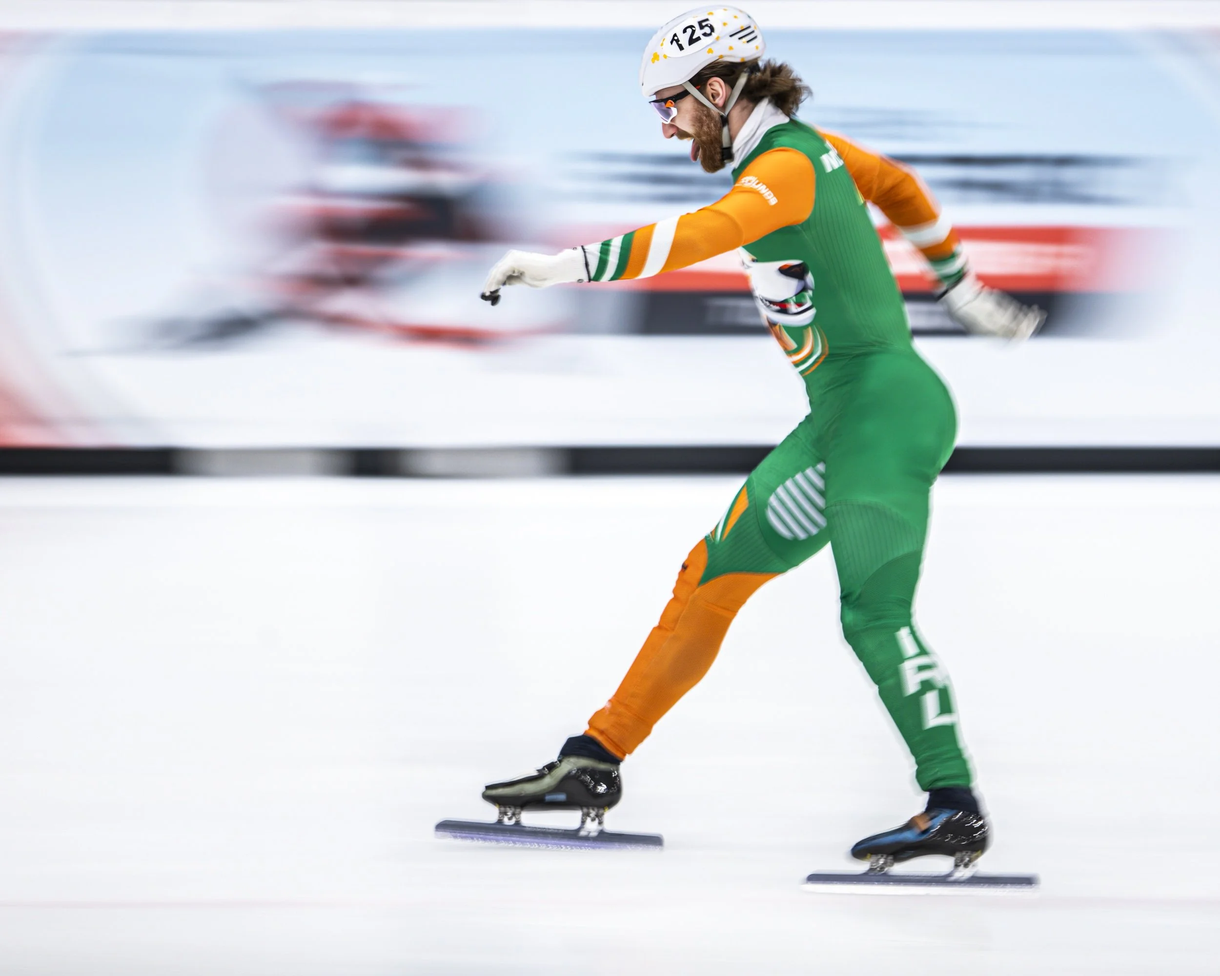 A male speed skater wearing a green and orange uniform, helmet, and sunglasses, racing on ice with blurred teammates in the background.