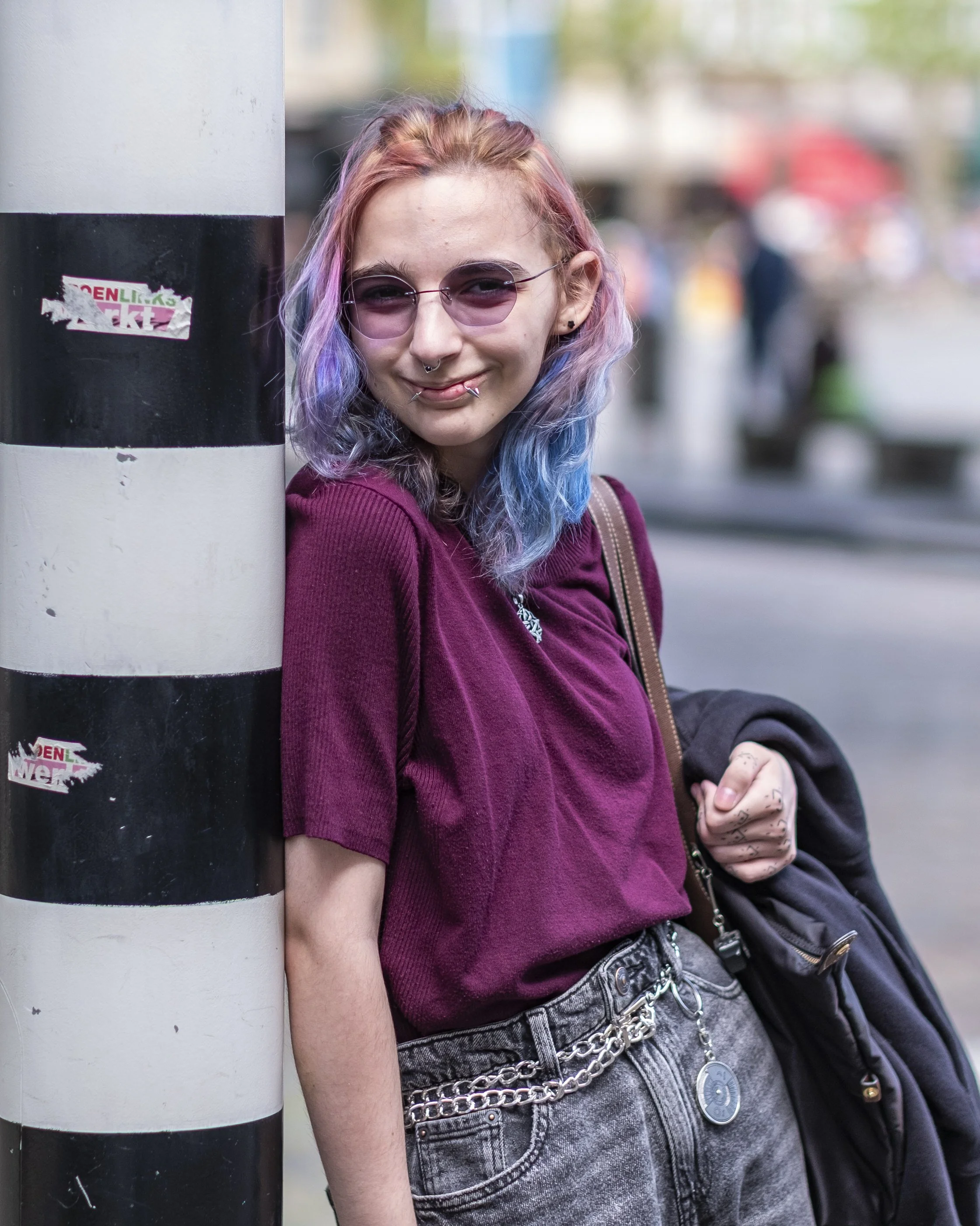 A young woman with multicolored hair, wearing sunglasses, a septum piercy, and a lip ring. She is leaning against a black-and-white striped pole on a city street, smiling at the camera. She has a chain belt and is carrying a black backpack.