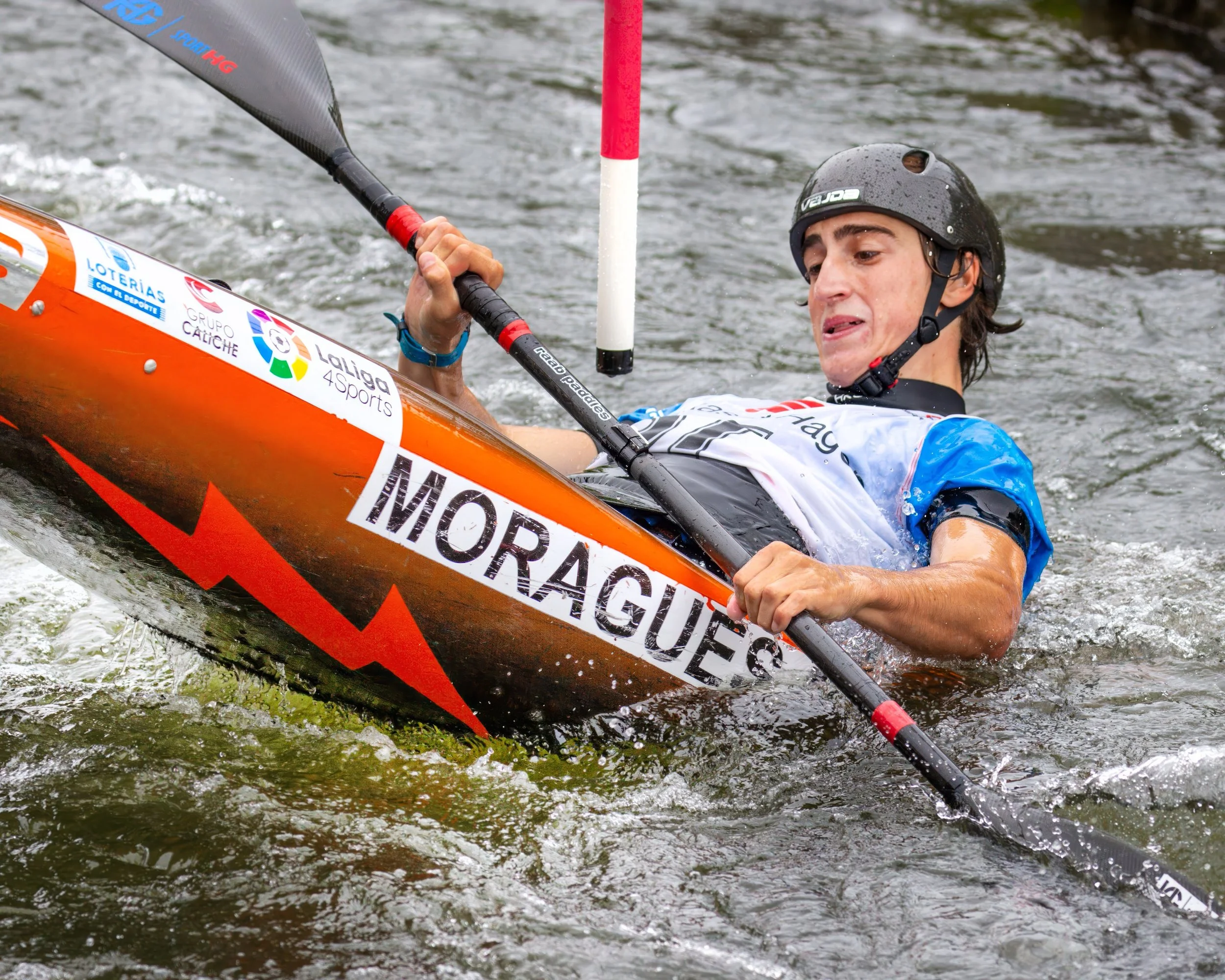 A kayaker in a helmet and sports gear navigates through water with a slalom gate pole in front of him, surrounded by turbulent water.