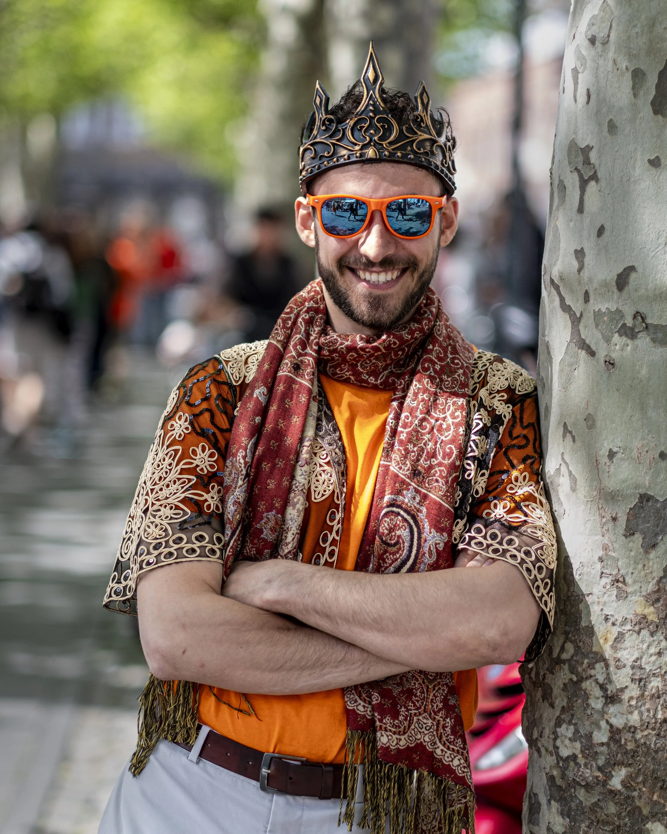 Man wearing orange sunglasses, a decorated crown, patterned jacket, and a scarf with crossed arms, standing next to a tree outdoors on a sunny day.