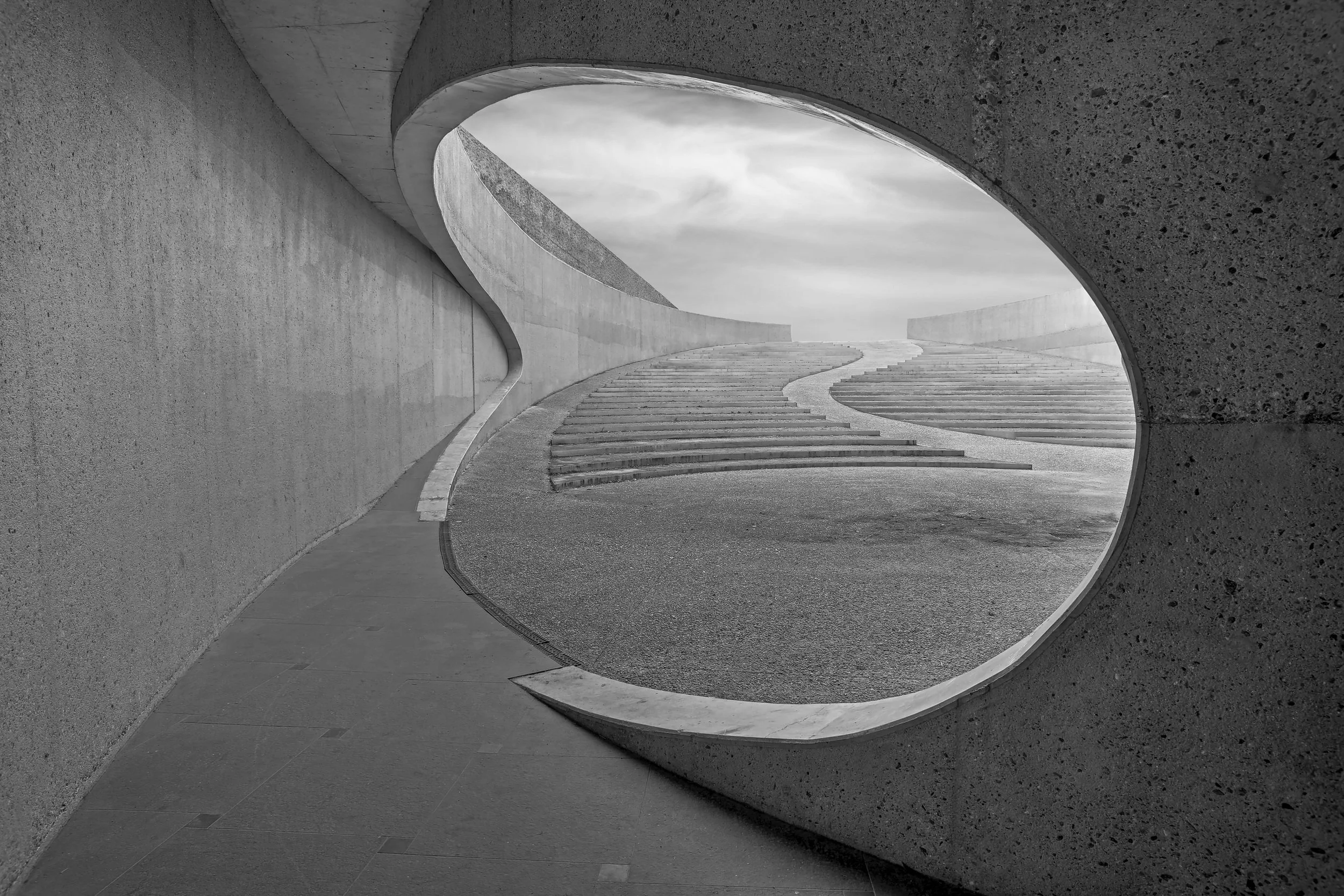 Black and white photo of a modern architectural structure with curved concrete walls, stairs, and an opening revealing the sky.