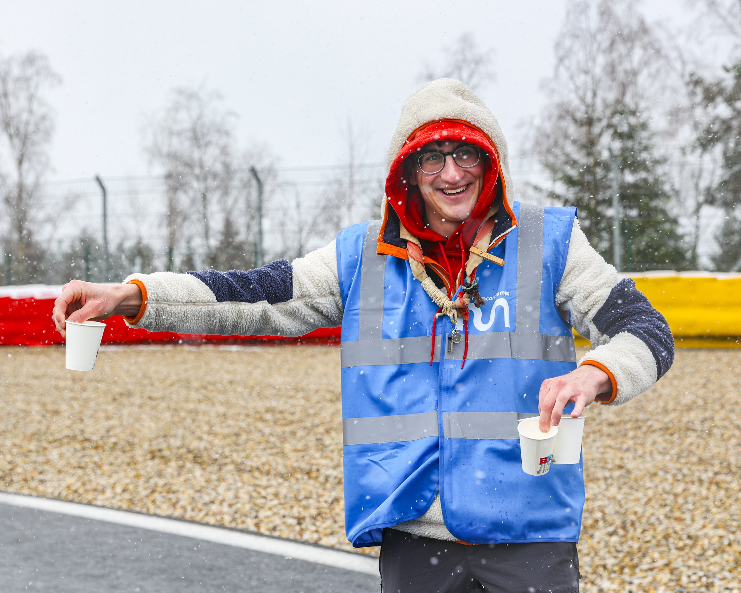 A man smiling outdoors in winter, wearing glasses, a beige beanie, a red hooded sweatshirt, and a blue safety vest. He is holding a white paper cup and has his arms outstretched as snow falls around him.