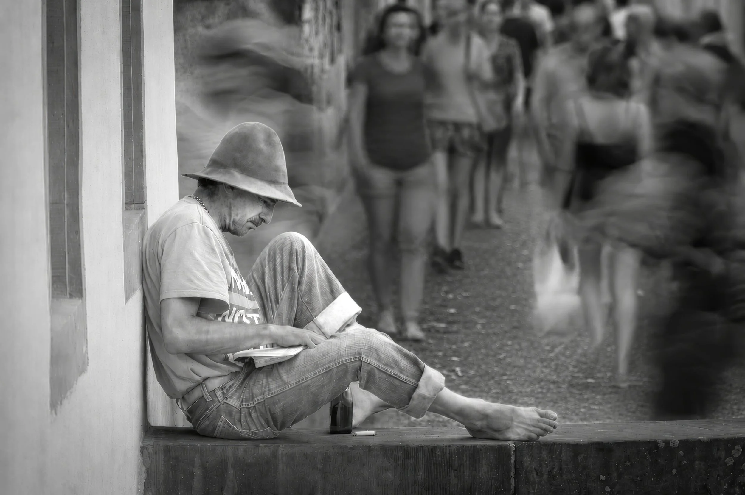 A man wearing a wide-brimmed straw hat and glasses, sitting against a wall, reading a book with other people in the background walking along a sidewalk.