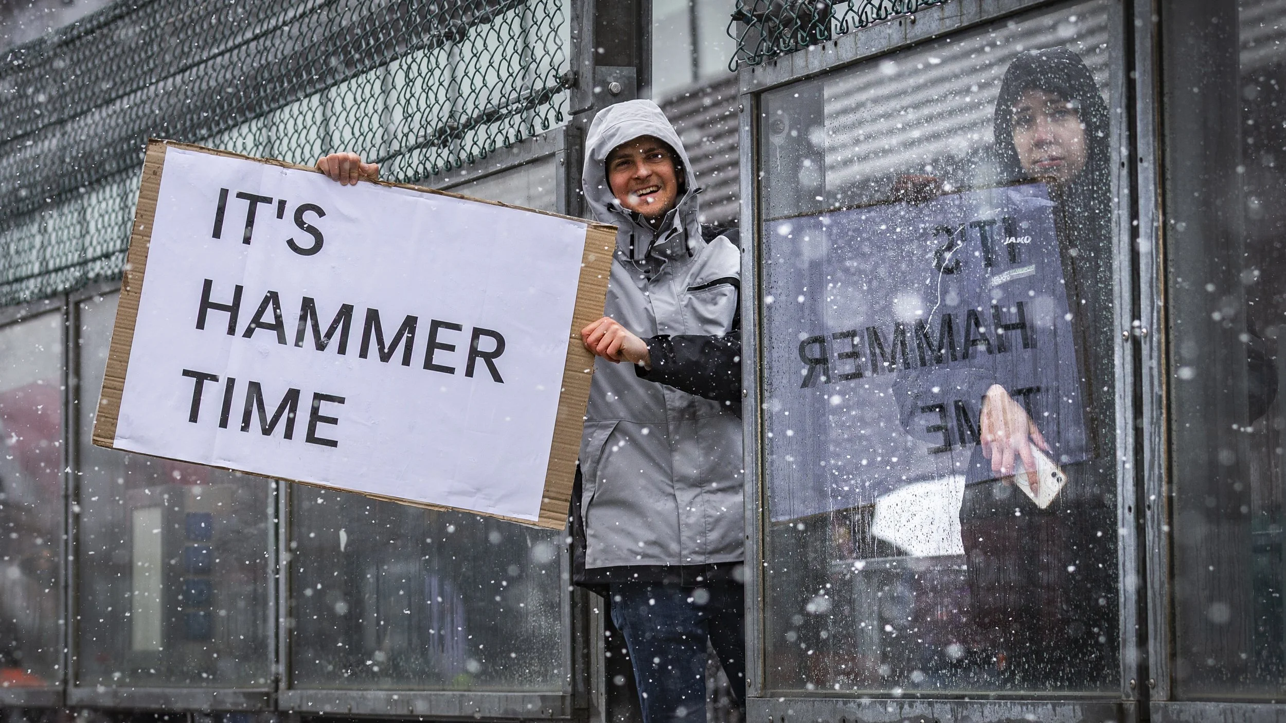 Two people standing at a bus stop in the snow; one holding a sign that says "IT'S HAMMER TIME" and the other looking out from behind a glass window.