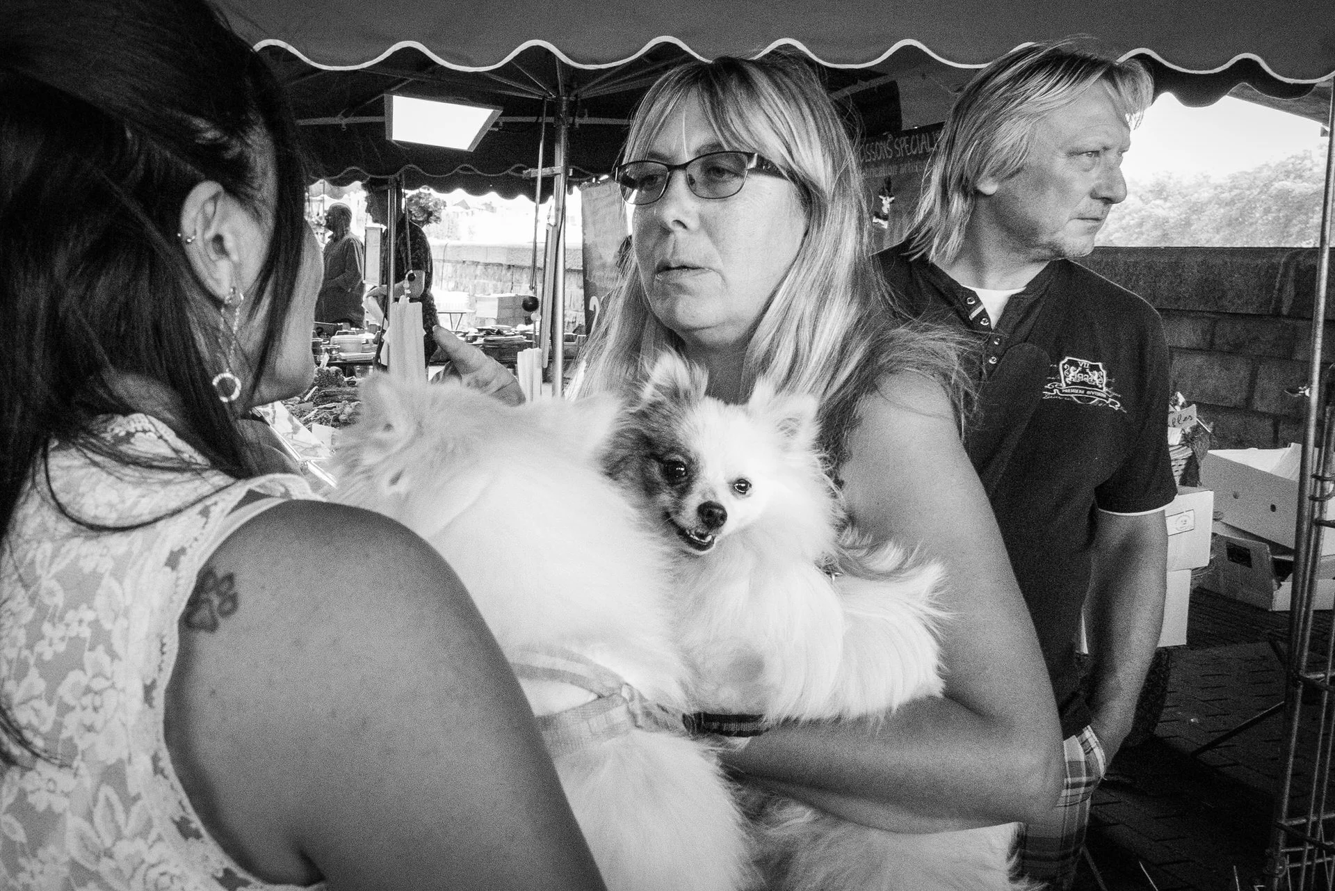 A woman holding a small fluffy dog, with a woman wearing glasses and a man standing nearby at an outdoor event under a tent.