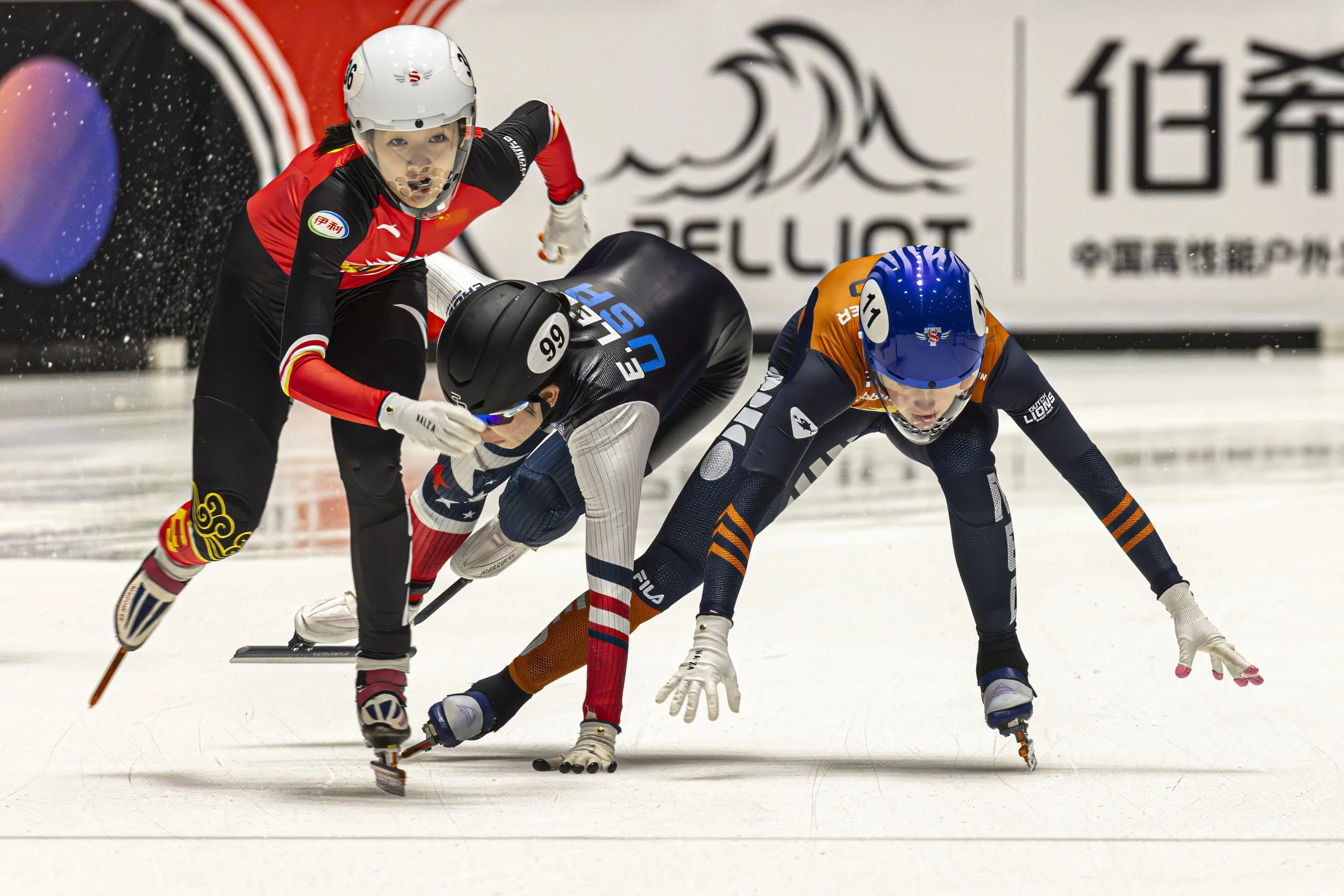 Two athletes in colorful uniforms competing in a speed skating race on ice, wearing helmets and skating gear, leaning forward on their skis.